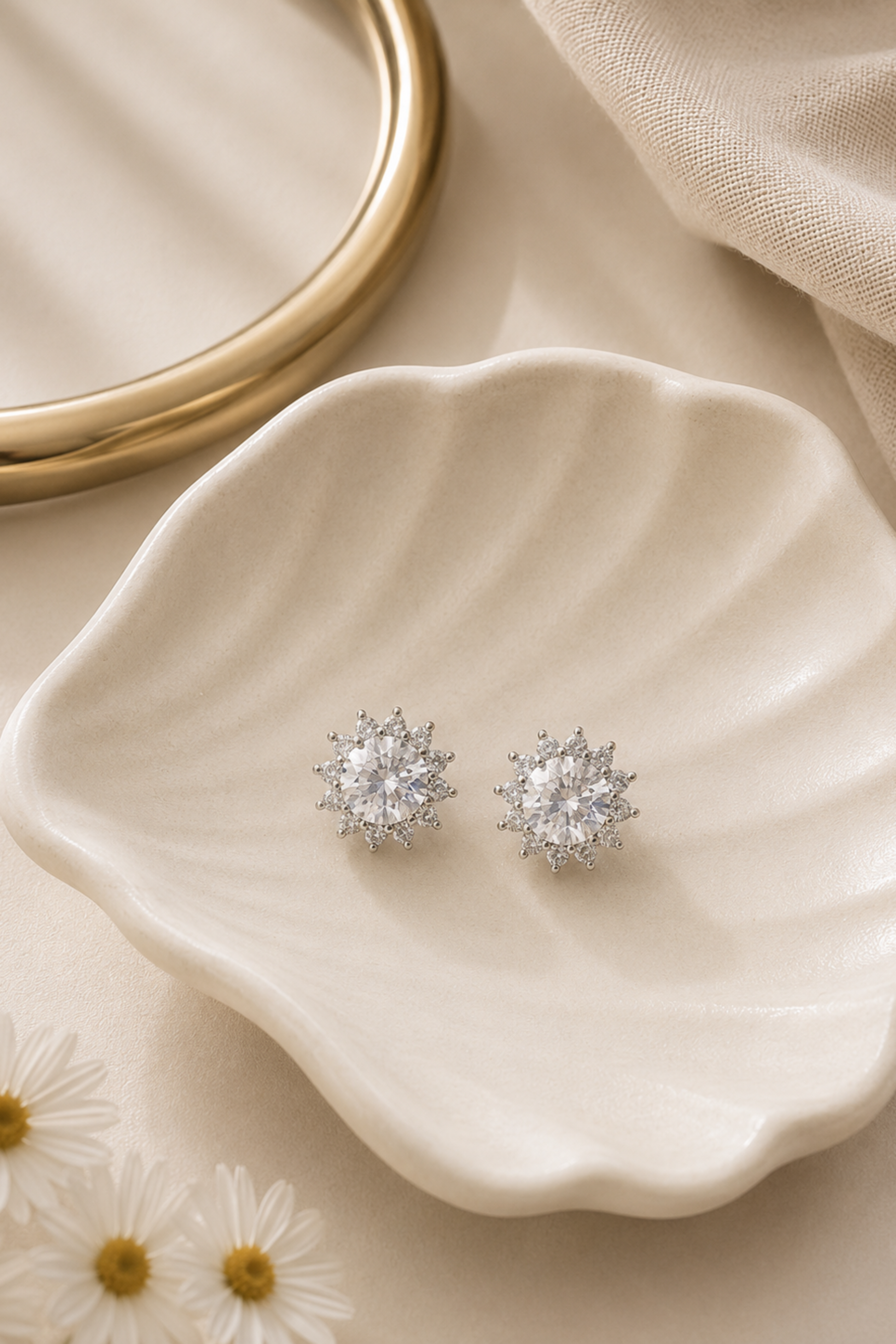A pair of silver-toned stud earrings with clear crystal clusters, displayed on a cream-colored scalloped dish with a gold ring and fabric in the background.