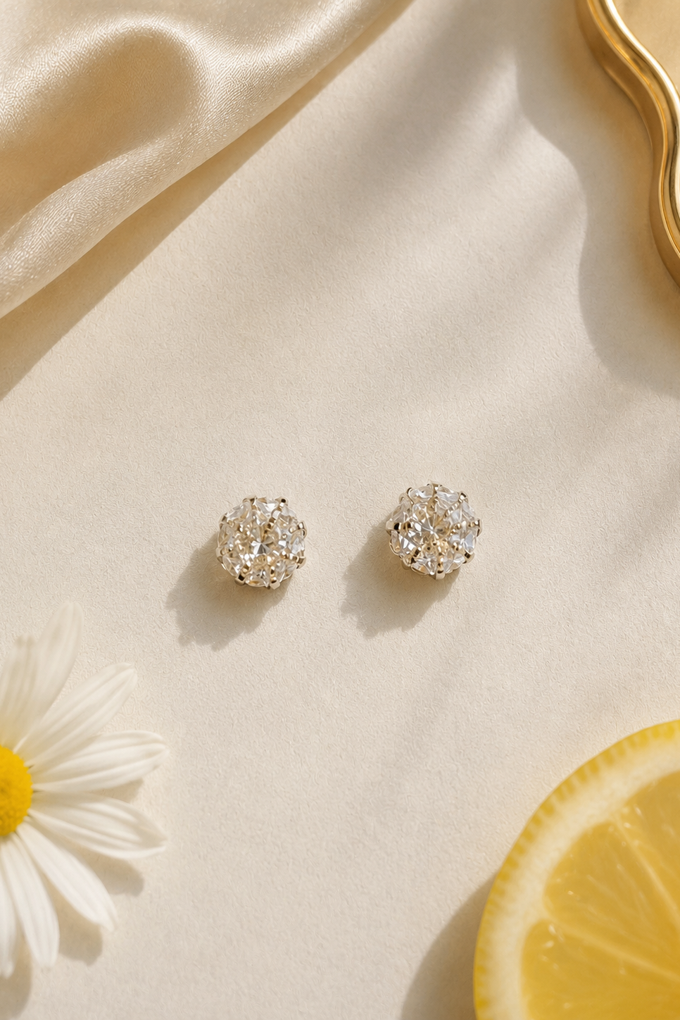 A pair of silver-toned stud earrings with clear gemstones, arranged on a cream satin surface with a daisy flower, a lemon slice, and a gold tray edge as styled props.