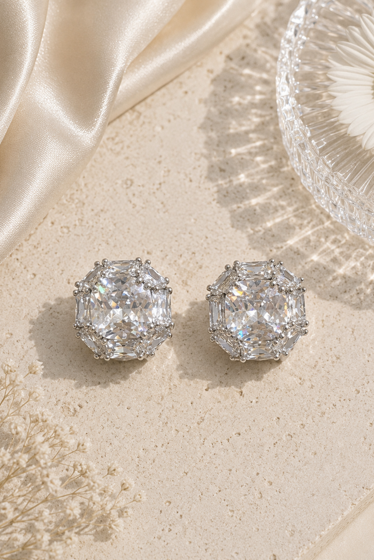 A pair of silver-toned stud earrings with large clear emerald-cut gemstones, displayed on a textured beige surface with cream satin fabric, dried flowers, and a glass bowl.