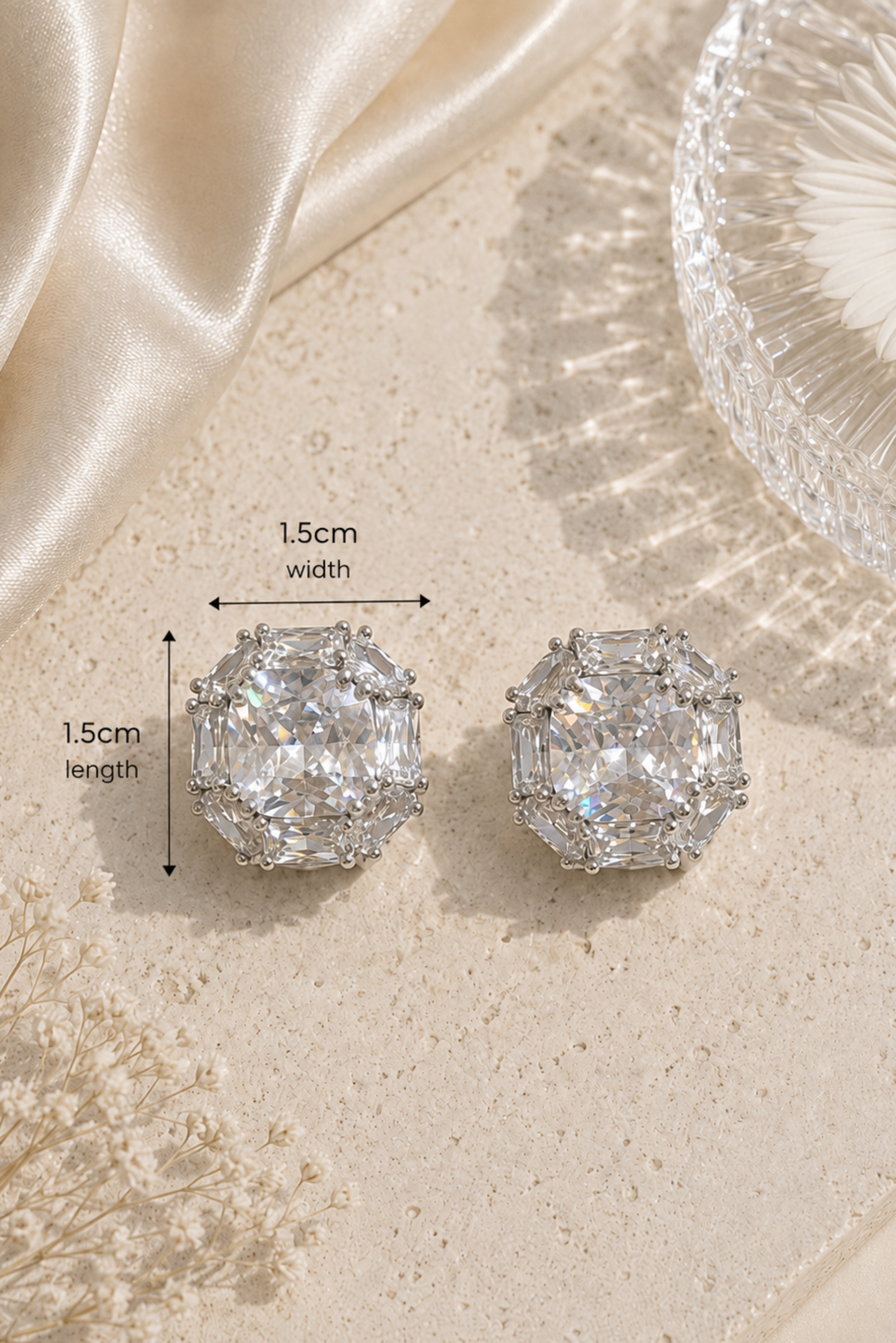 A pair of silver-toned stud earrings with large clear emerald-cut stones, shown on a beige textured surface with satin fabric, dried flowers, and a glass bowl. Black measurement arrows and text indicate 1.