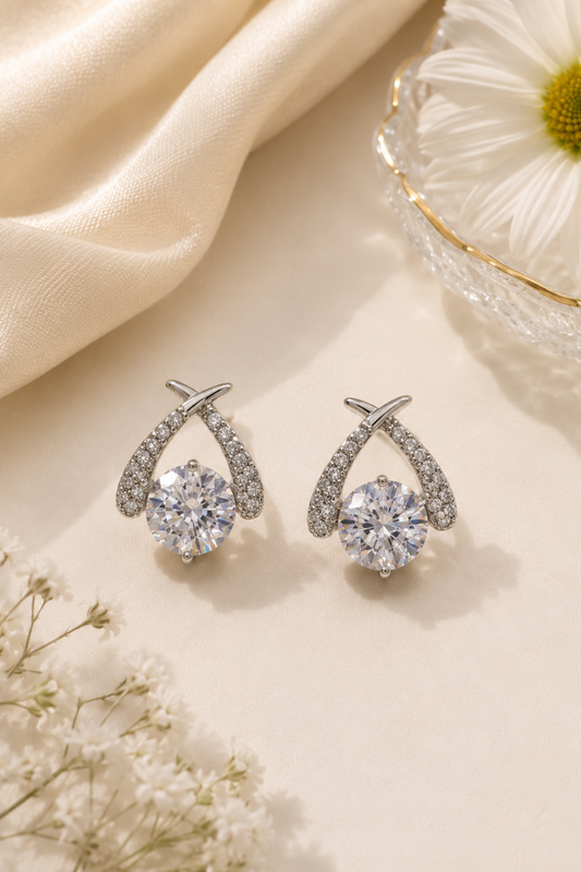 A pair of silver-toned teardrop-shaped earrings with clear round gemstones, styled on a cream surface with a satin fabric, a daisy flower, and baby's breath.