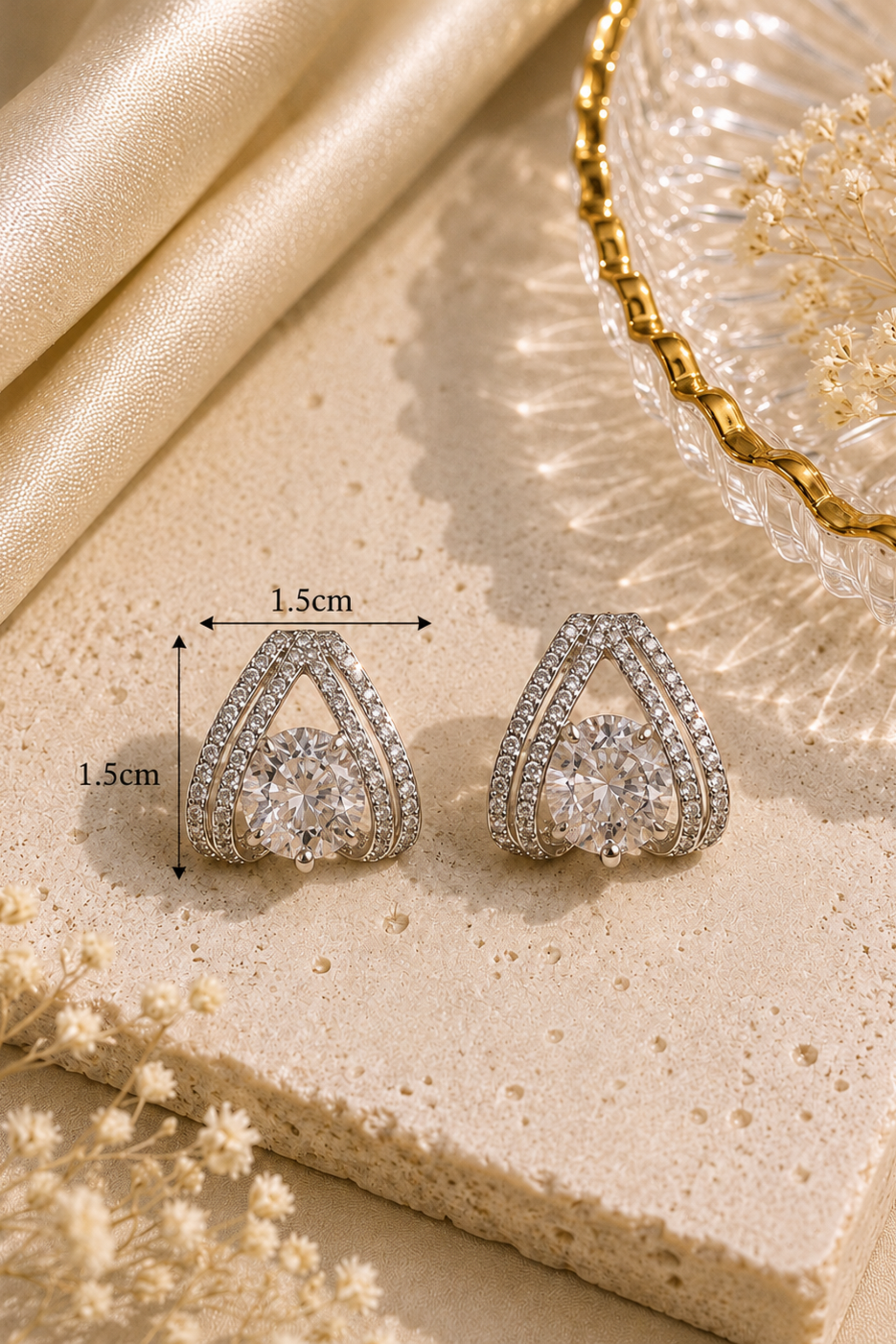 A pair of silver-toned teardrop-shaped stud earrings with clear gemstones, displayed on a beige stone slab with dimension arrows showing 1.5cm width and 1.