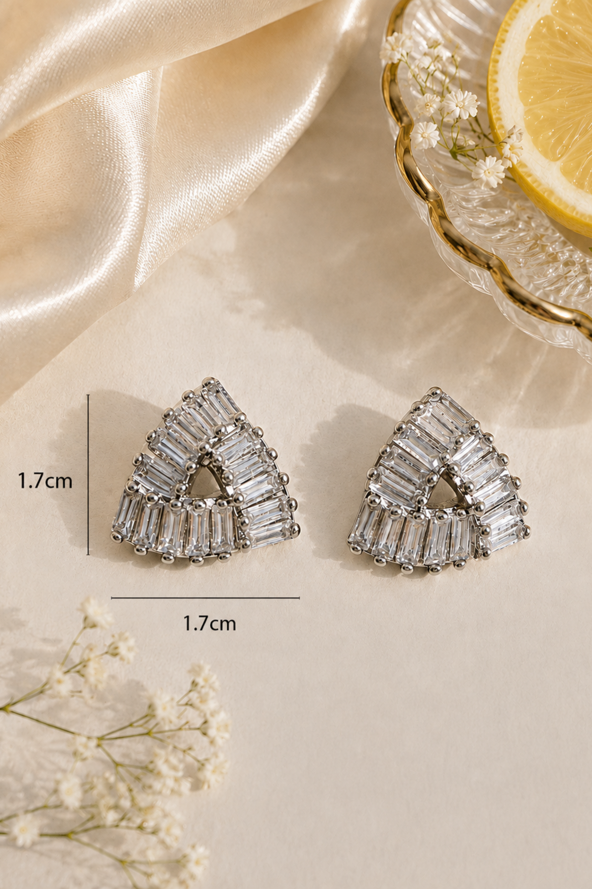 A pair of silver-toned triangle stud earrings with baguette-cut clear stones, shown on a cream surface with satin fabric, small white flowers, and a lemon slice in a glass dish. Measurement lines and text indicate 1.