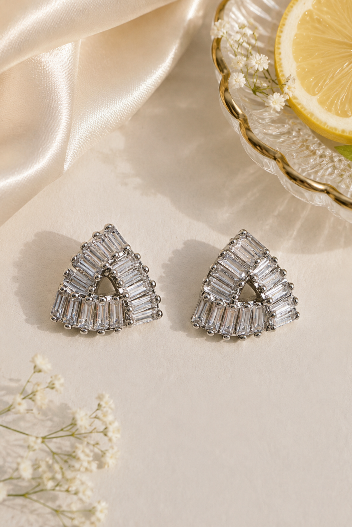 A pair of silver-toned triangular stud earrings with baguette-cut clear stones, styled on a cream surface with satin fabric, small white flowers, and a lemon slice in a glass dish.