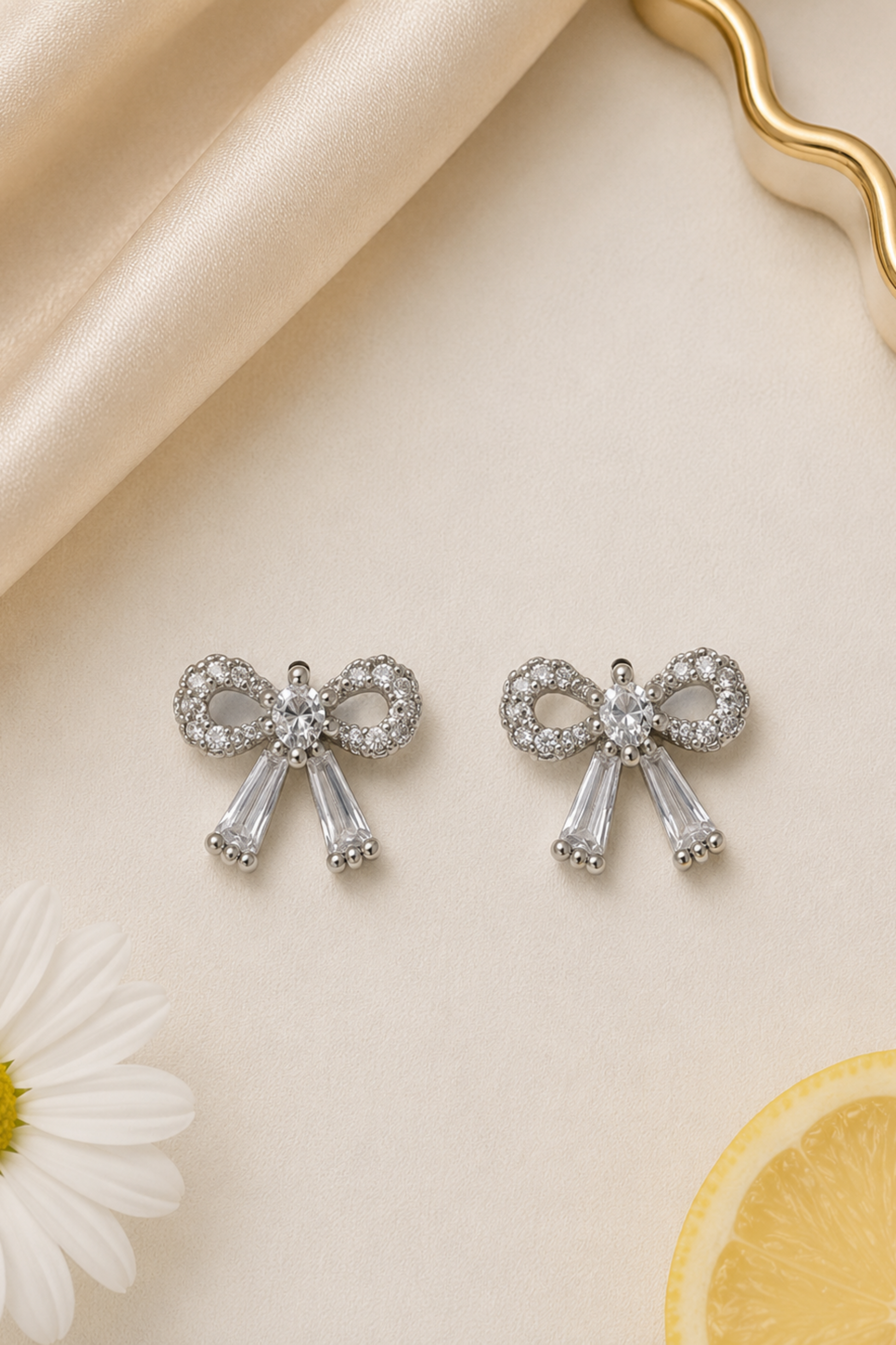 A pair of silver bow-shaped stud earrings adorned with clear crystals, displayed on a light beige textured surface with a cream satin fabric, a white daisy, and a lemon slice.
