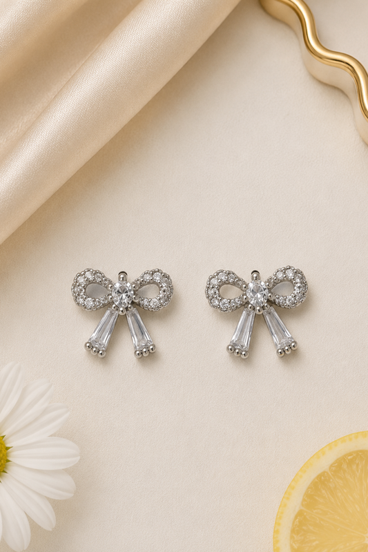 A pair of silver bow-shaped stud earrings adorned with clear crystals, displayed on a light beige textured surface with a cream satin fabric, a white daisy, and a lemon slice.