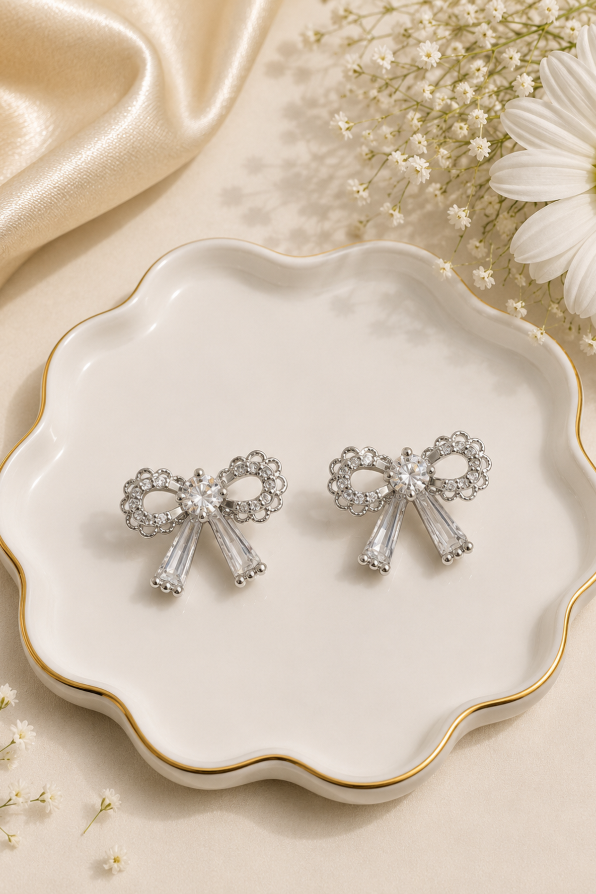 A pair of silver bow-shaped stud earrings with clear rhinestone accents and faceted clear drops, displayed on a scalloped white ceramic dish with gold trim, surrounded by satin fabric, baby's breath flowers, and a white daisy.