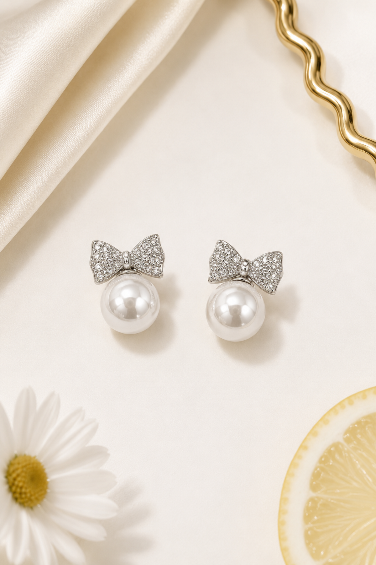 A pair of silver bow stud earrings with clear pavé crystals and a white pearl drop, styled on a cream background with a daisy, a lemon slice, and cream satin fabric.