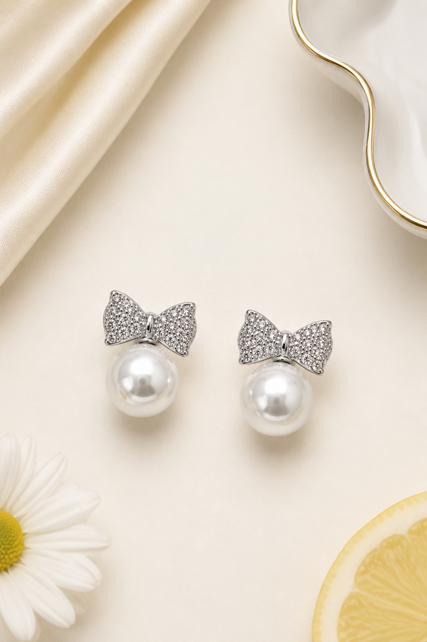 A pair of silver bow stud earrings with clear pavé detailing and a white pearl drop, styled on a cream background with satin, a daisy, and a lemon slice.