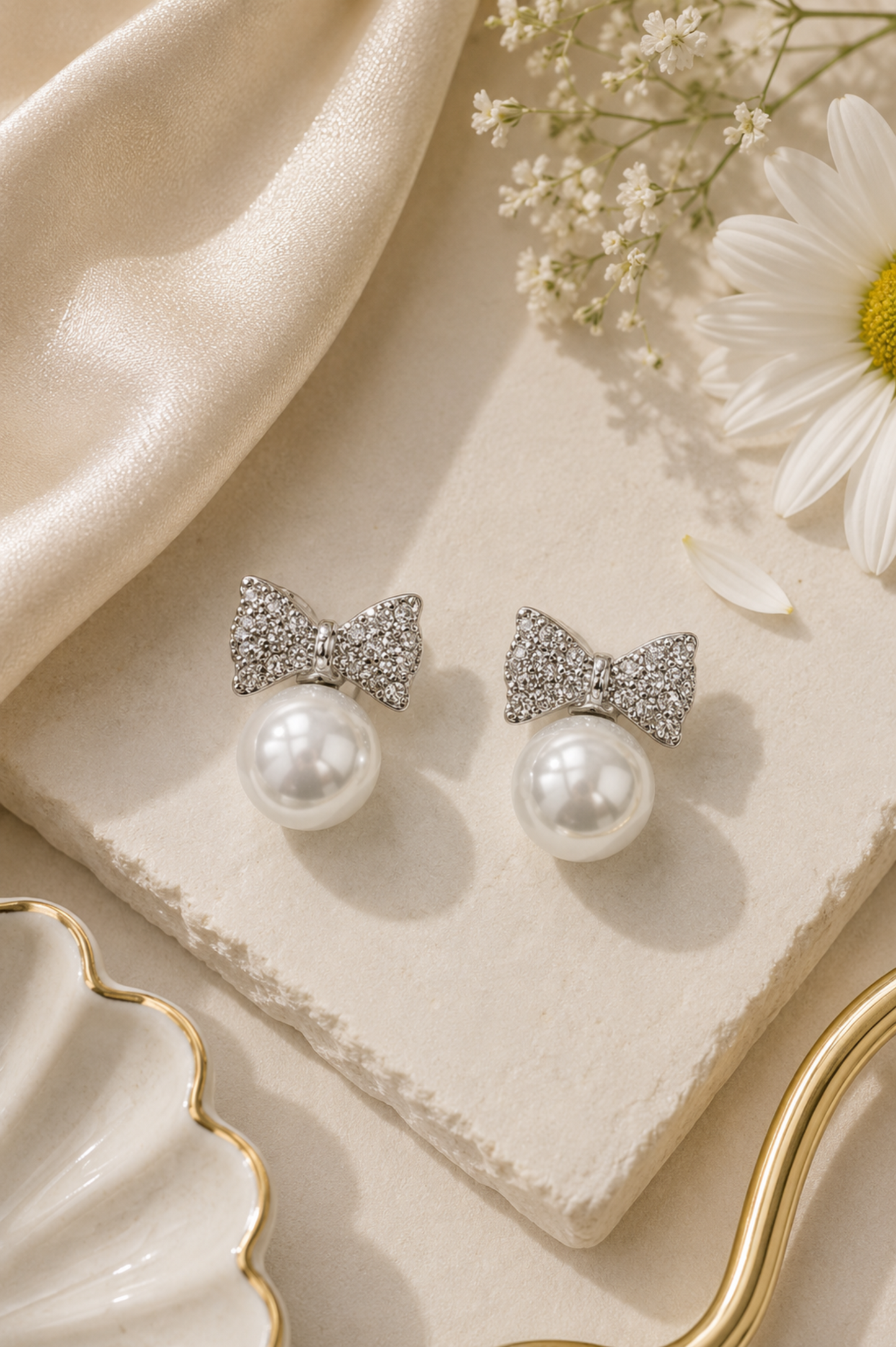 A pair of silver bow stud earrings with sparkling pavé crystal tops and white faux pearls below, displayed on a cream stone slab with a cream satin cloth, a daisy flower, and a gold-rimmed dish.
