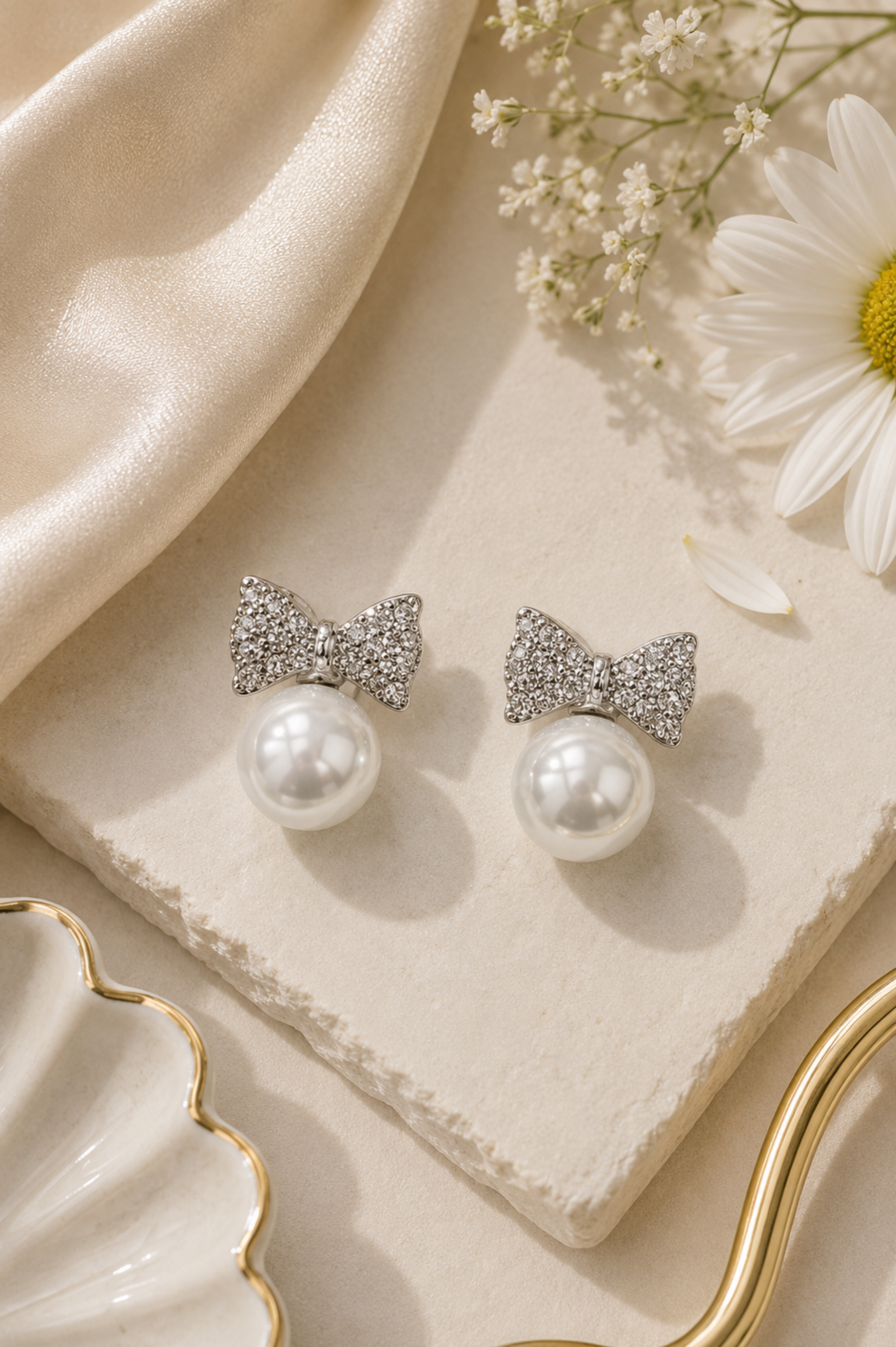 A pair of silver bow stud earrings with sparkling pavé crystal tops and white faux pearls below, displayed on a cream stone slab with a cream satin cloth, a daisy flower, and a gold-rimmed dish.
