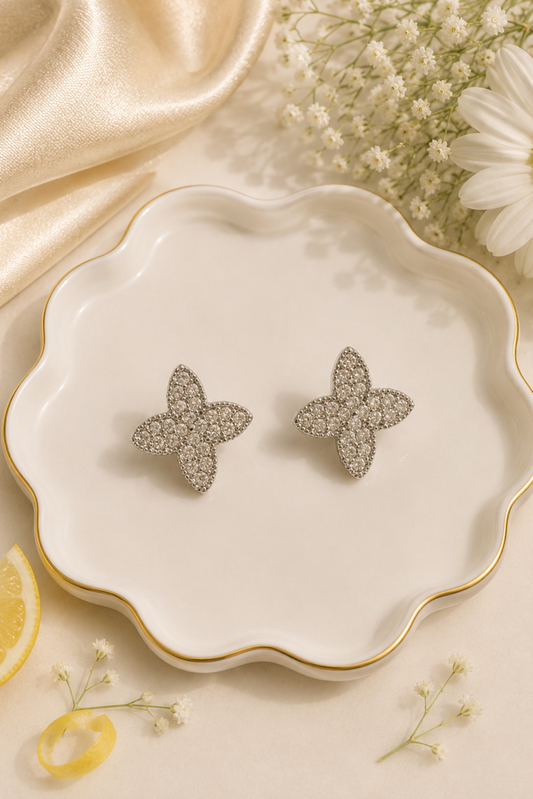 A pair of silver butterfly-shaped stud earrings covered in clear crystals, displayed on a white scalloped ceramic dish with gold trim, surrounded by decorative props including a daisy, baby's breath, a lemon slice, and a gold satin cloth.