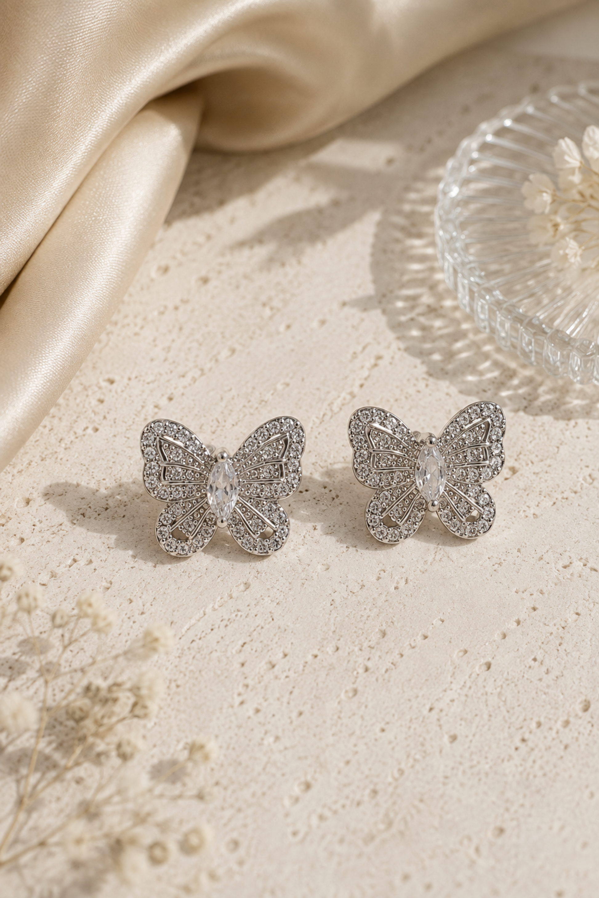 A pair of silver butterfly stud earrings adorned with clear crystals, displayed on a textured beige surface with a draped satin fabric and a glass dish in the background.