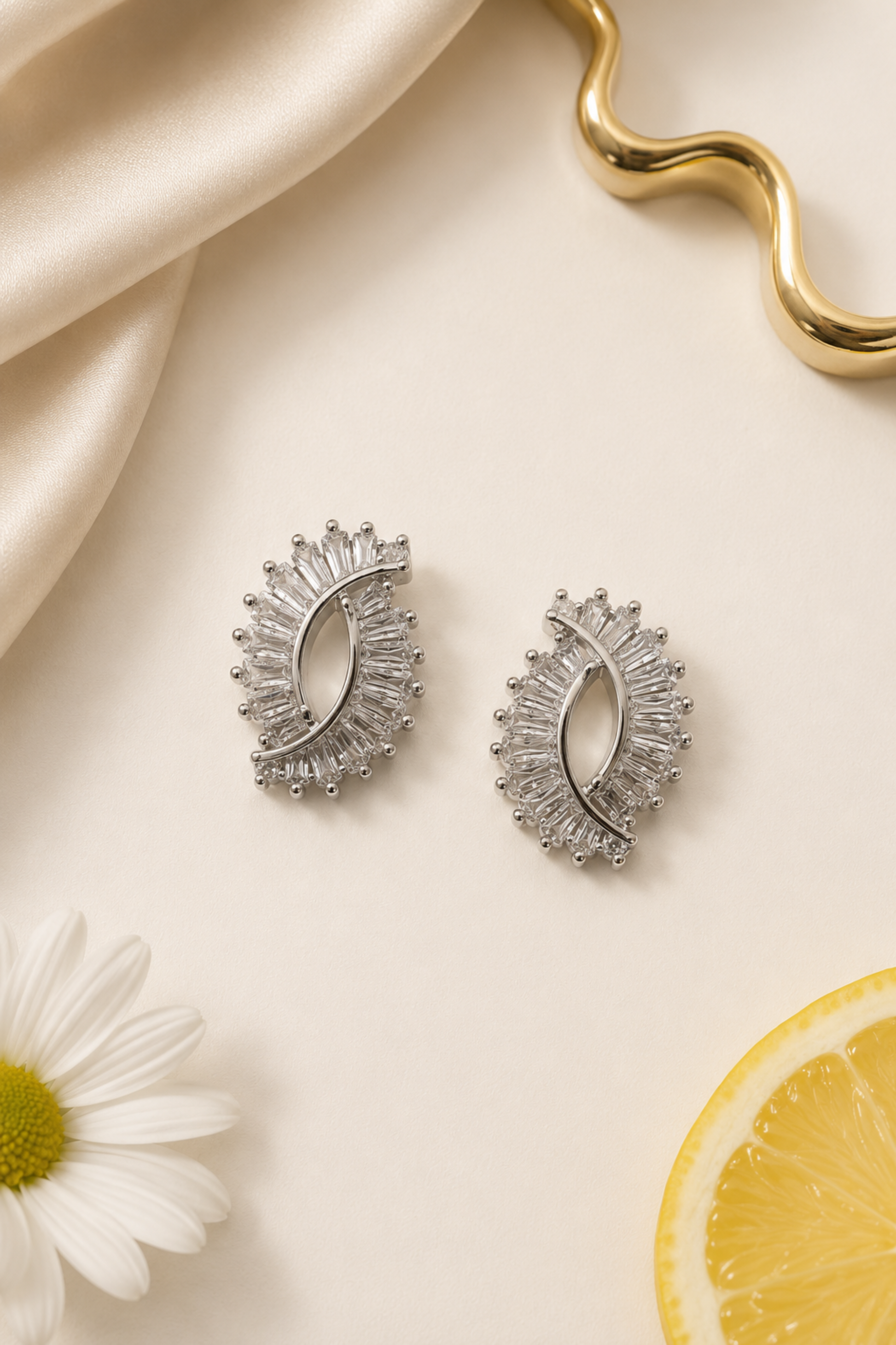 A pair of silver crescent-shaped stud earrings adorned with clear baguette and round crystals, styled on a cream background with a white daisy flower, a lemon slice, and a gold wavy object.