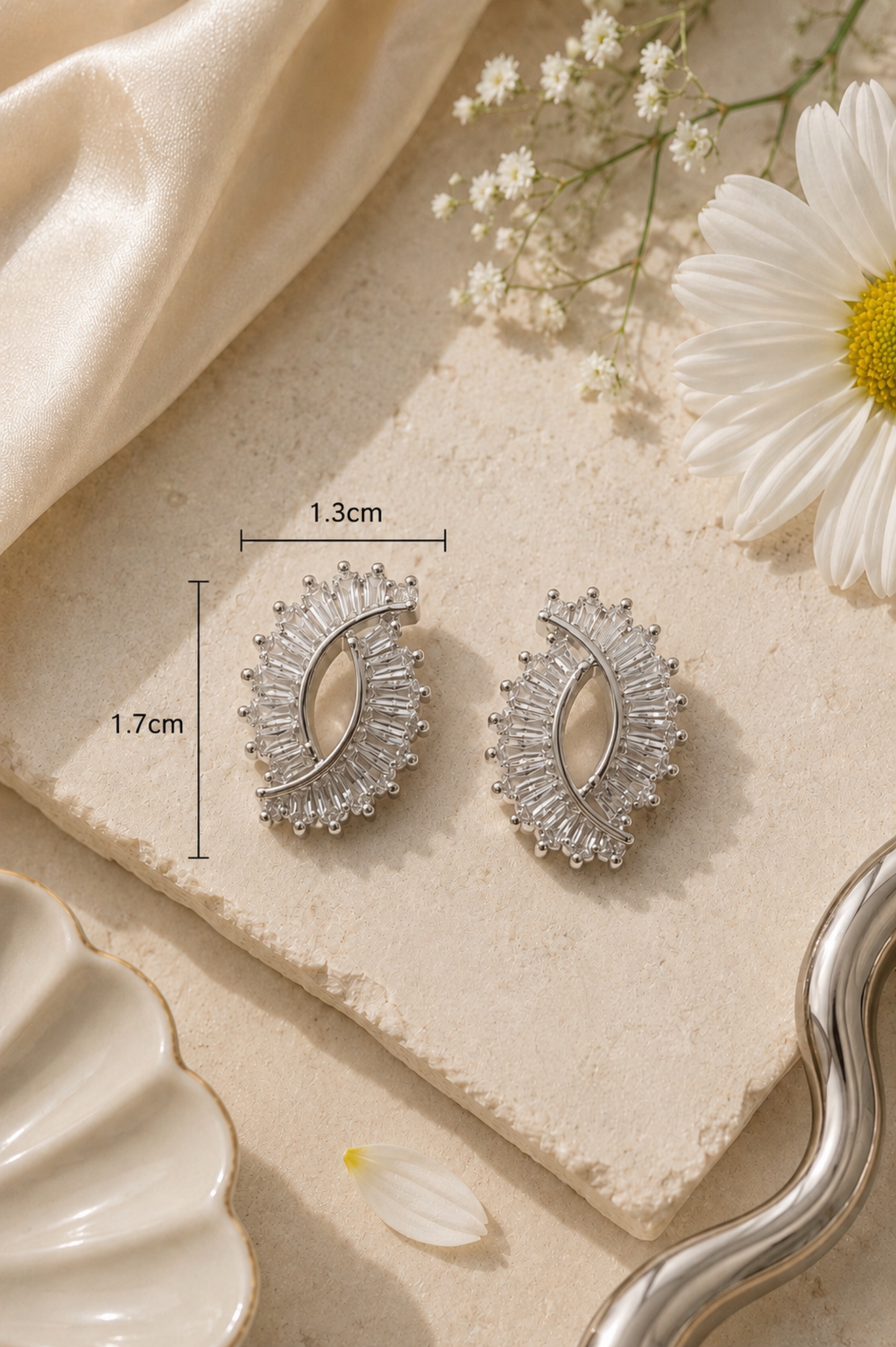 A pair of silver crescent-shaped stud earrings with clear crystal detailing, displayed on a textured beige stone slab with dimension lines showing 1.3cm width and 1.