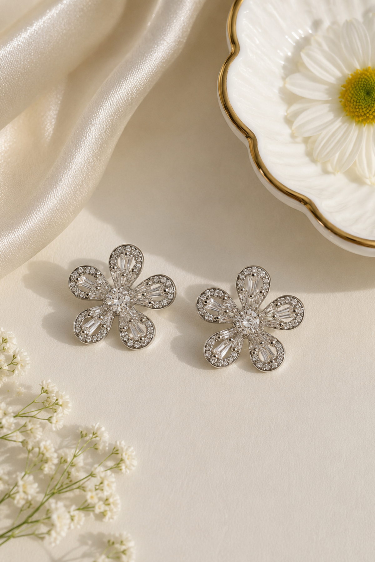 A pair of silver flower-shaped stud earrings adorned with clear crystals, arranged on a cream-colored surface with decorative satin fabric, a white daisy in a dish, and small white flowers.