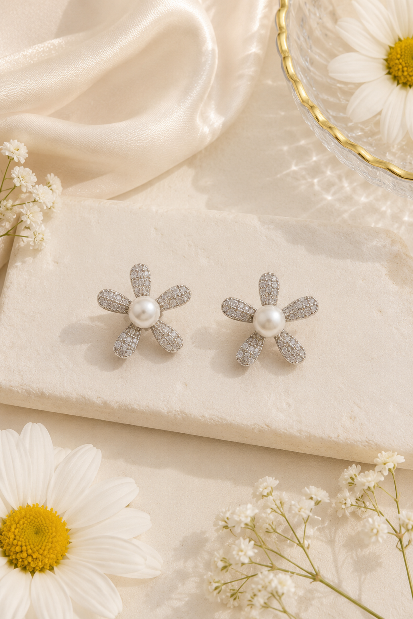 A pair of silver flower-shaped stud earrings with a central white pearl and pavé-set accents, displayed on a cream stone slab with white daisies and satin fabric.