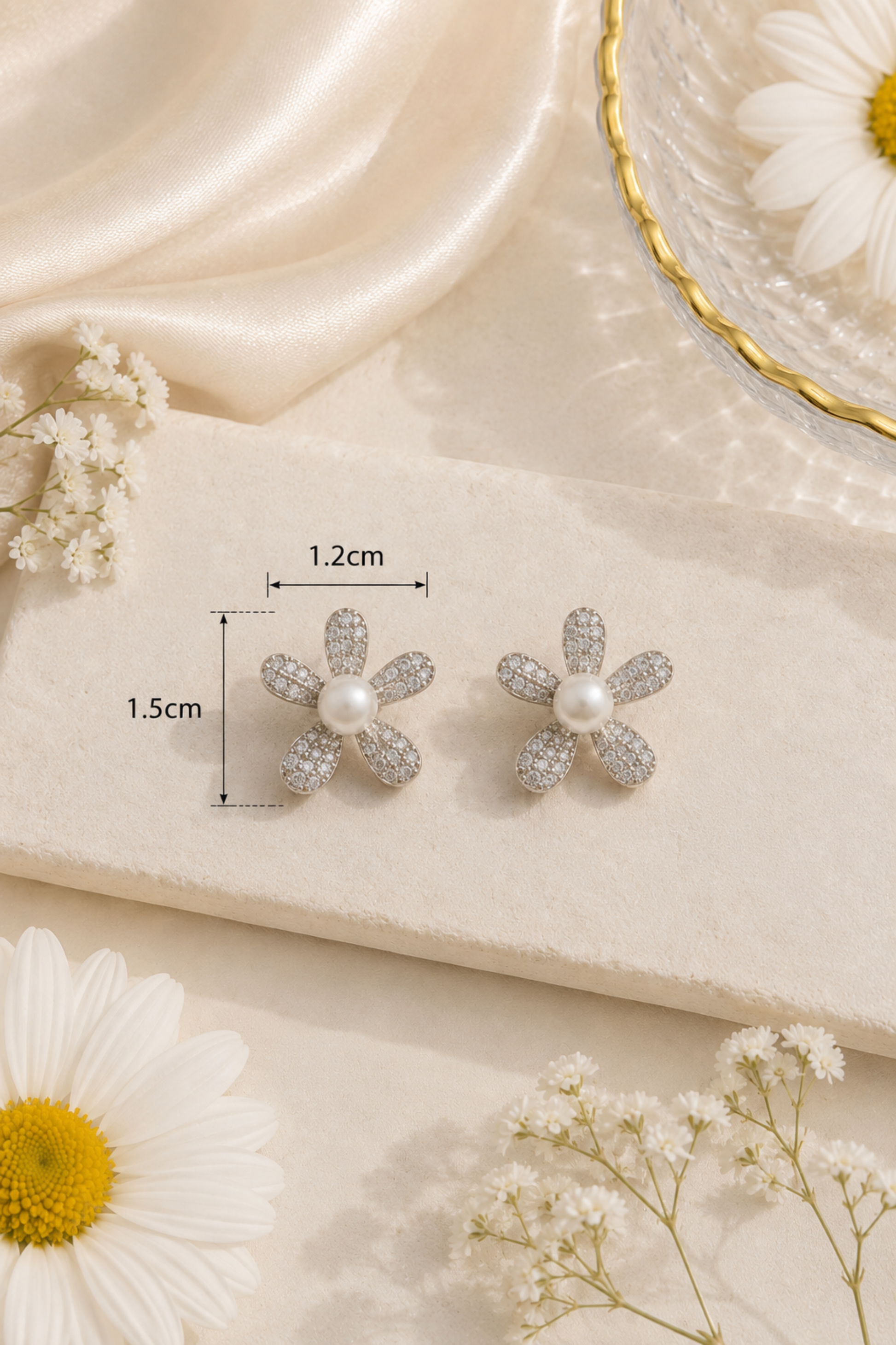 A pair of silver flower stud earrings with clear pavé details and a central white pearl, displayed on a cream fabric block with dimension lines showing 1.2cm width and 1.