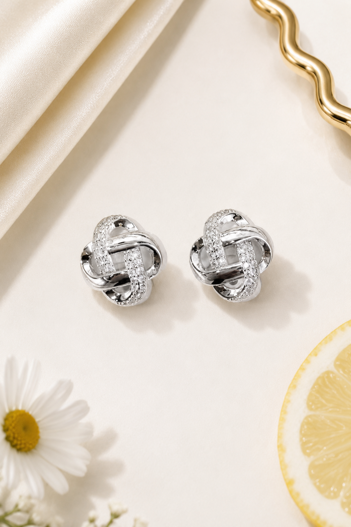 A pair of silver knot-shaped stud earrings with clear pavé stones, displayed on a cream-colored surface with a daisy, a lemon slice, and elegant fabric props.