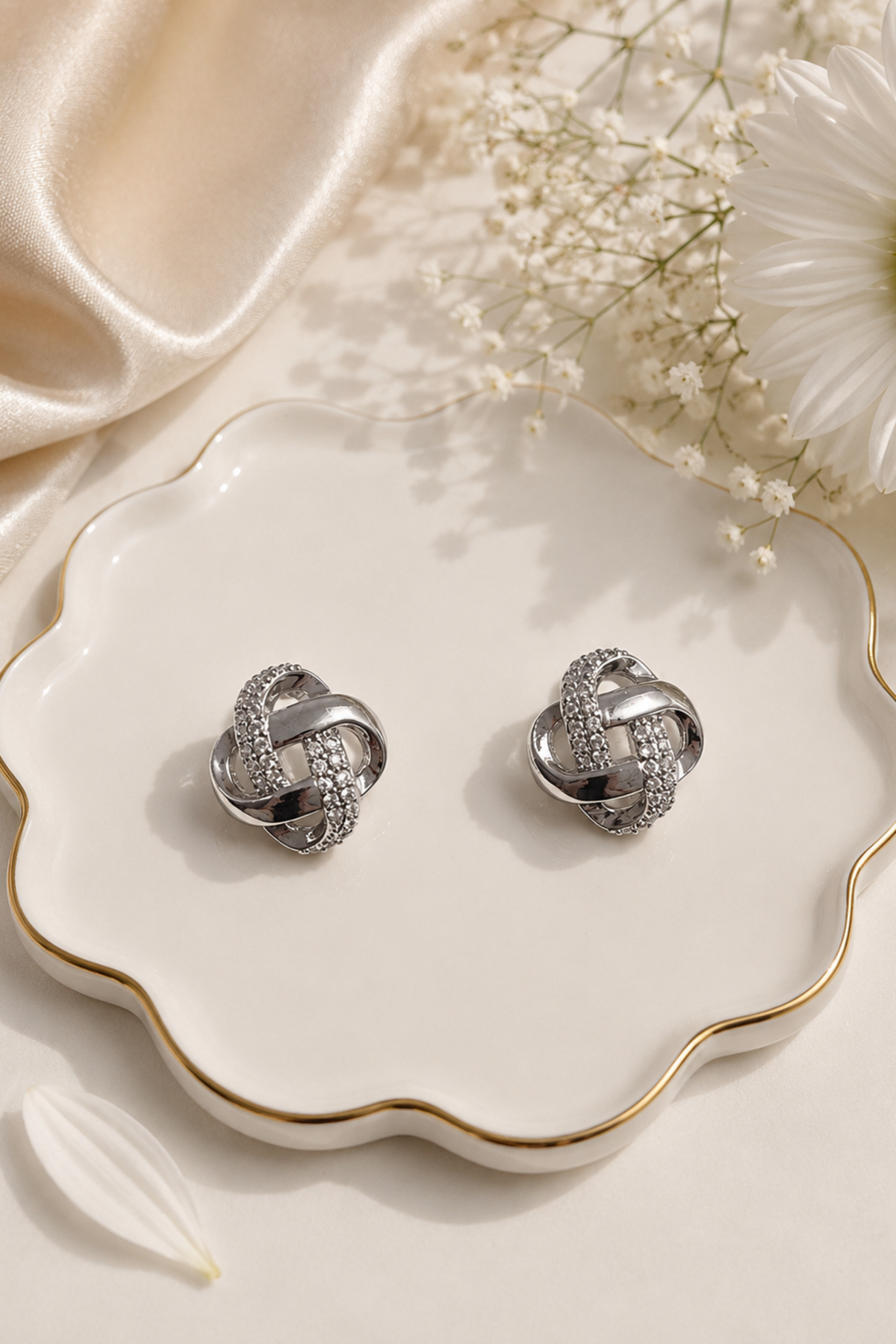 A pair of silver knot-shaped stud earrings with pavé-set clear stones, displayed on a scalloped white ceramic dish with gold trim, styled with satin fabric, baby's breath, and a white daisy.