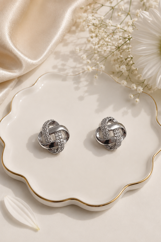 A pair of silver knot-shaped stud earrings with pavé-set clear stones, displayed on a scalloped white ceramic dish with gold trim, styled with satin fabric, baby's breath, and a white daisy.
