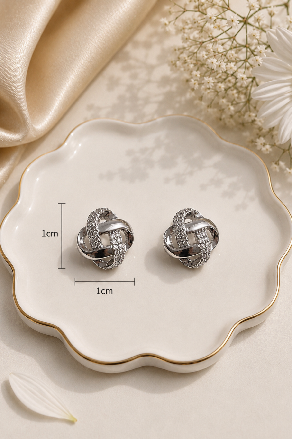 A pair of silver knot-style stud earrings displayed on a scalloped cream dish with gold trim, surrounded by satin fabric, baby's breath flowers, and a white daisy. Measurement markers indicate approximately 1 cm height and 1 cm width.
