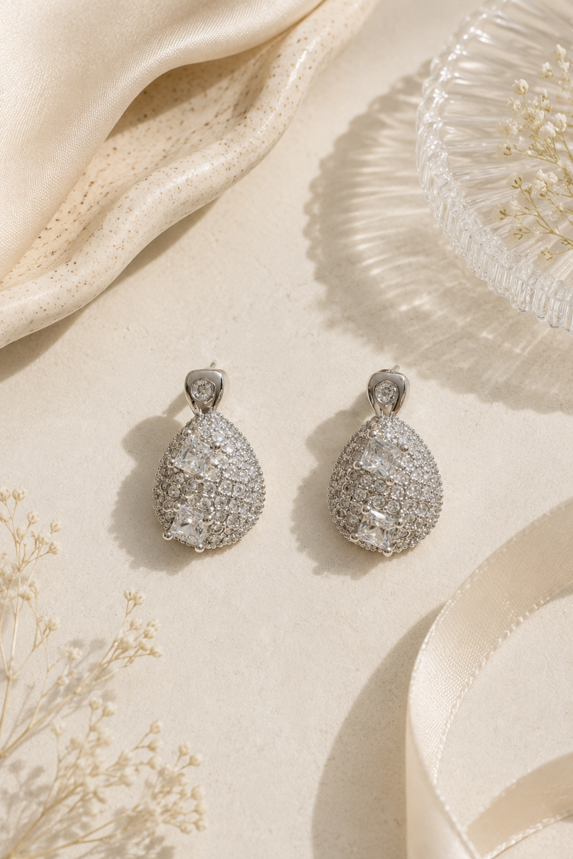 A pair of silver teardrop-shaped drop earrings covered in sparkling clear pavé stones, displayed on a cream-colored surface with draped satin fabric, ribbon, and dried flowers.