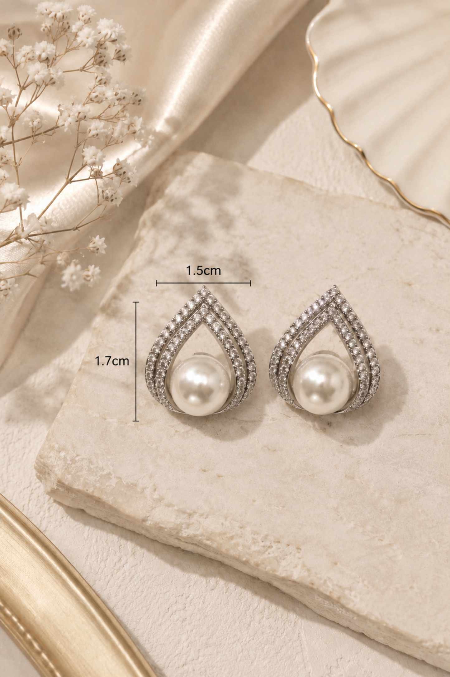 A pair of silver teardrop-shaped drop earrings with white pearl centers and small clear stones, displayed on a textured cream stone with size markers showing 1.5cm width and 1.