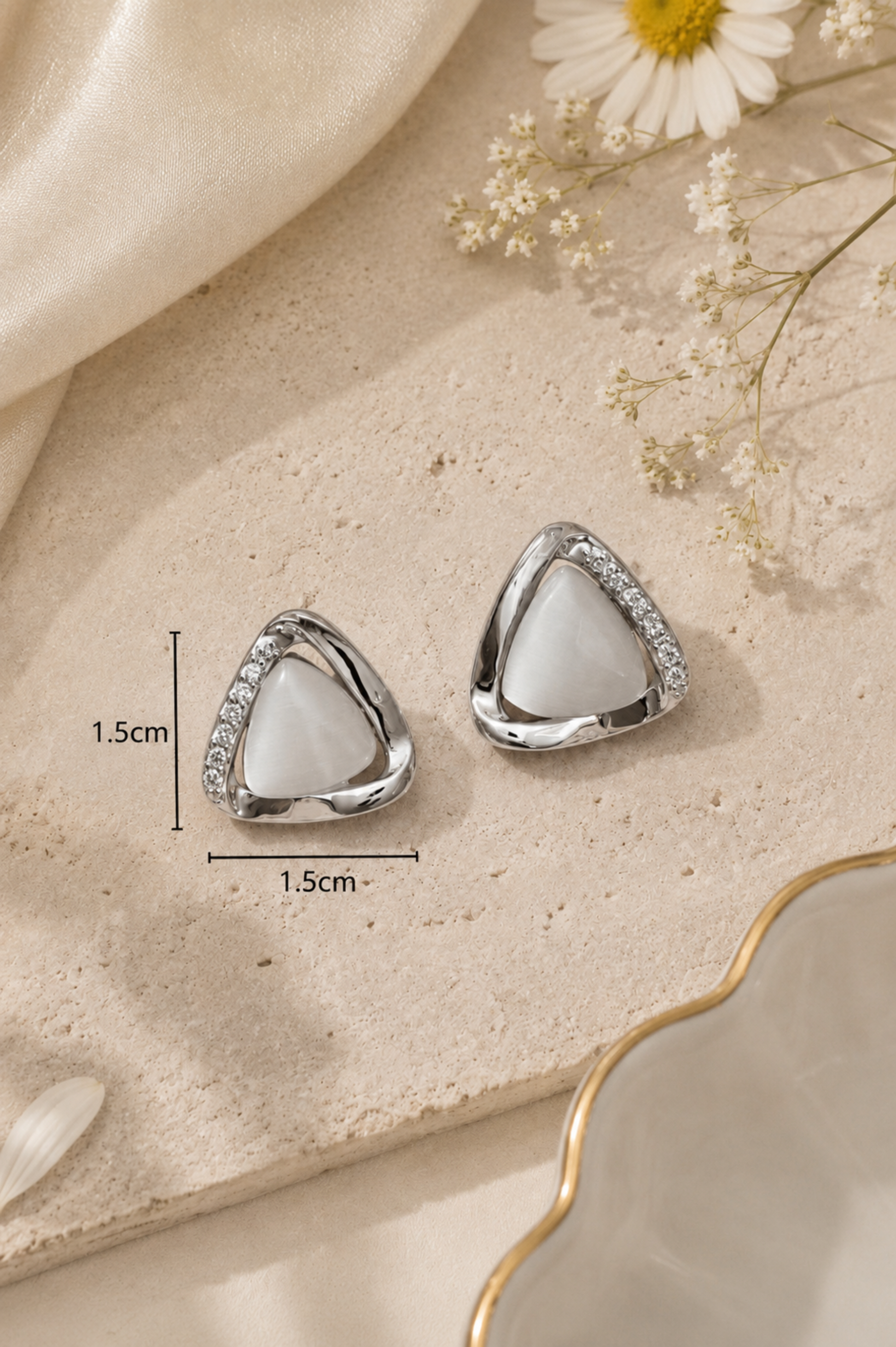 A pair of silver triangle stud earrings with white inlay and small clear stones along the edges, displayed on a beige textured surface with measurement annotations showing 1.5cm by 1.