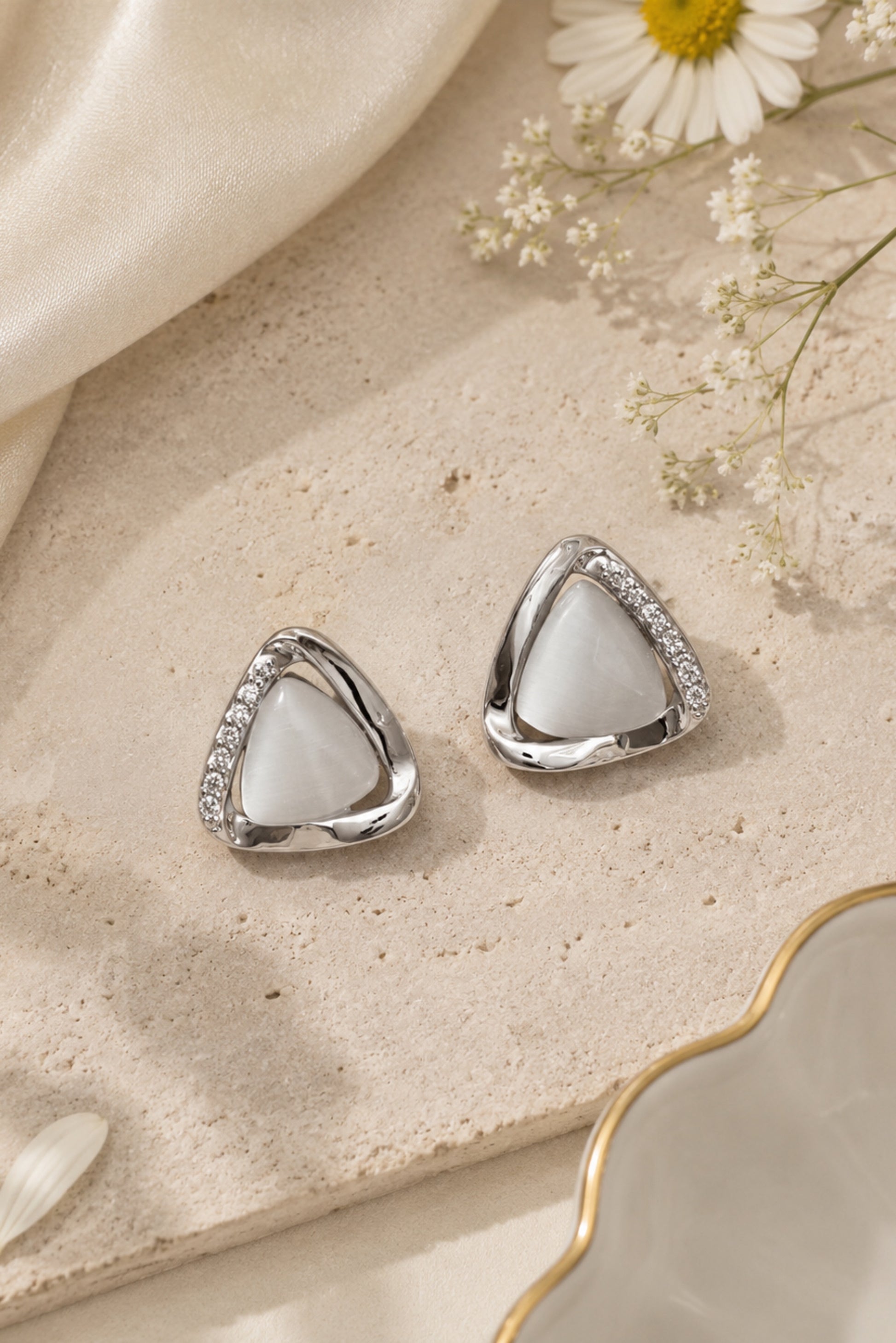 A pair of silver triangle stud earrings with white mother-of-pearl centers and small clear stones along the edges, styled on a textured beige surface with cream satin, dried flowers, and a gold-rimmed dish.