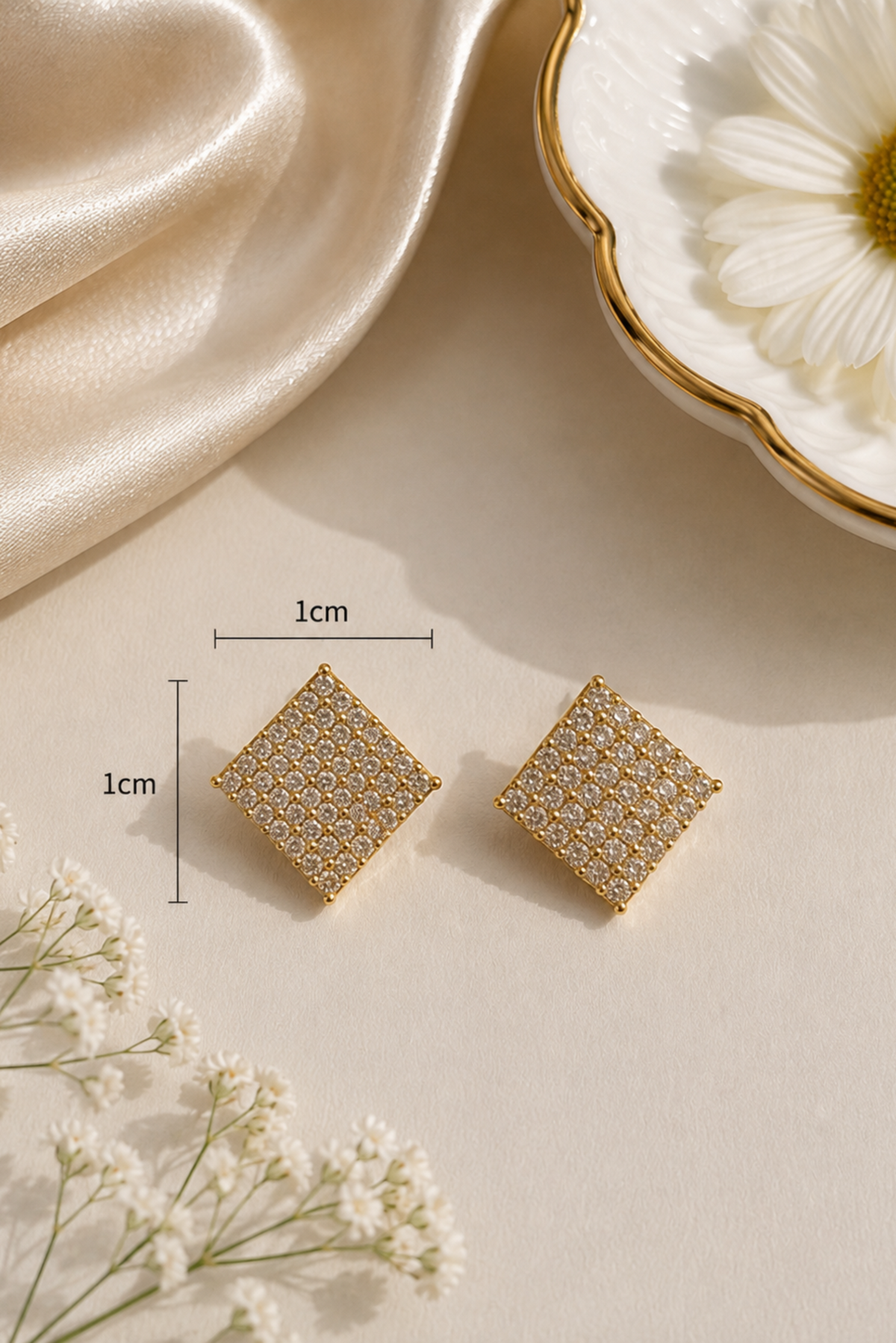 A pair of small square gold-toned earrings with a textured honeycomb pattern, displayed on a beige surface with dimension lines indicating 1cm by 1cm. The scene includes a cream satin cloth, a white daisy in a gold-rimmed dish, and small white flowers.