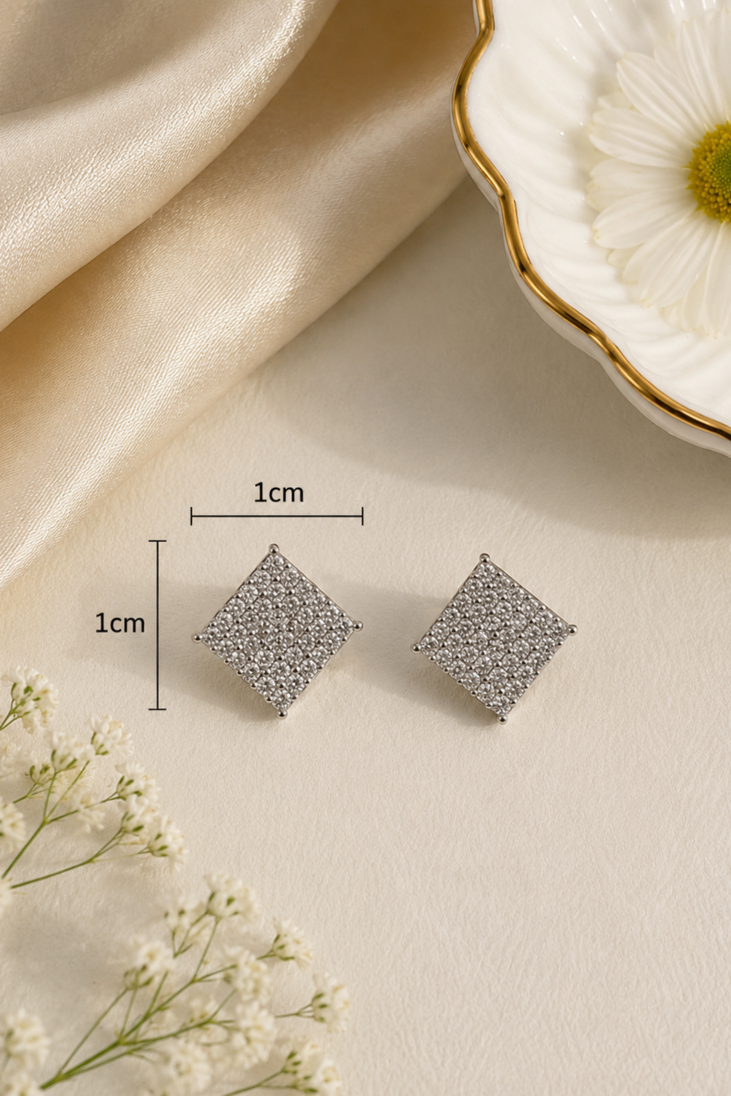 A pair of small square stud earrings with a textured silver pattern, displayed on a beige surface with dimension lines indicating a 1cm by 1cm size. The background includes draped champagne-colored satin fabric, white baby's breath flowers, and a white flower dish with gold trim.