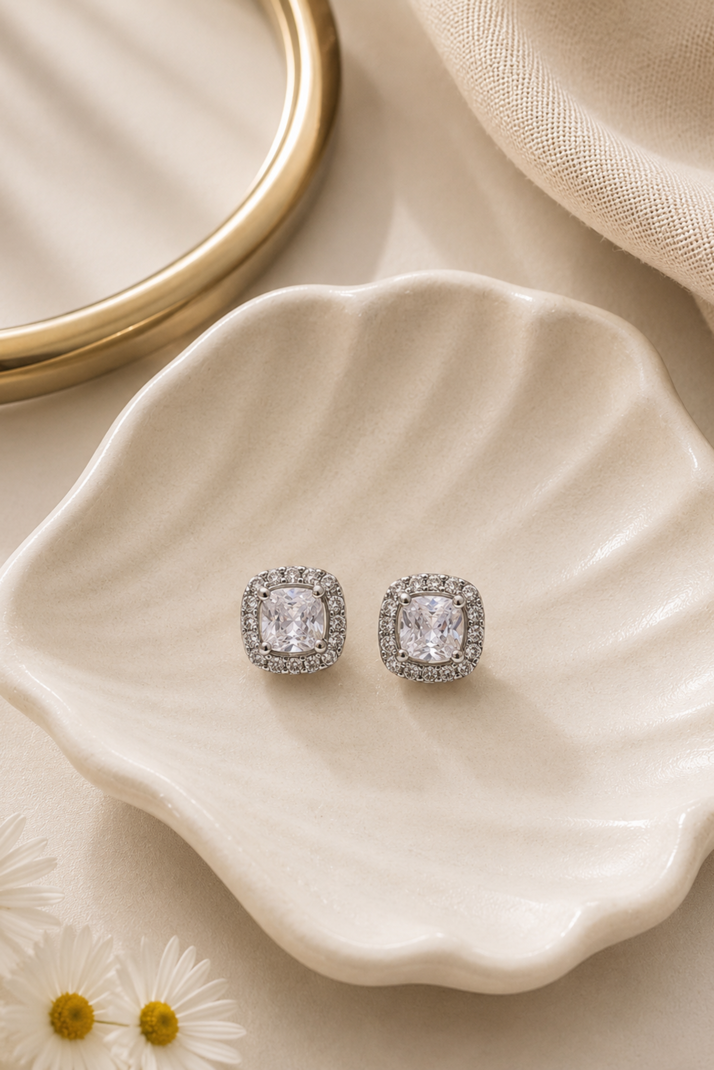A pair of square cushion-cut stud earrings with clear stones in silver settings, displayed on a scalloped cream ceramic dish with a gold ring and beige fabric in the background.