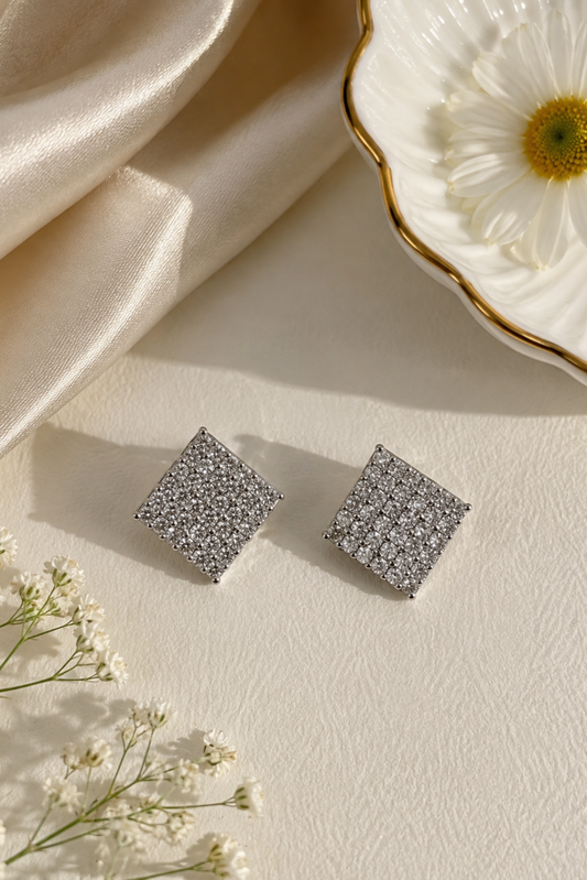 A pair of square stud earrings covered in small clear stones, displayed on a cream textured surface with draped satin fabric, a white daisy in a gold-rimmed dish, and small white flowers.