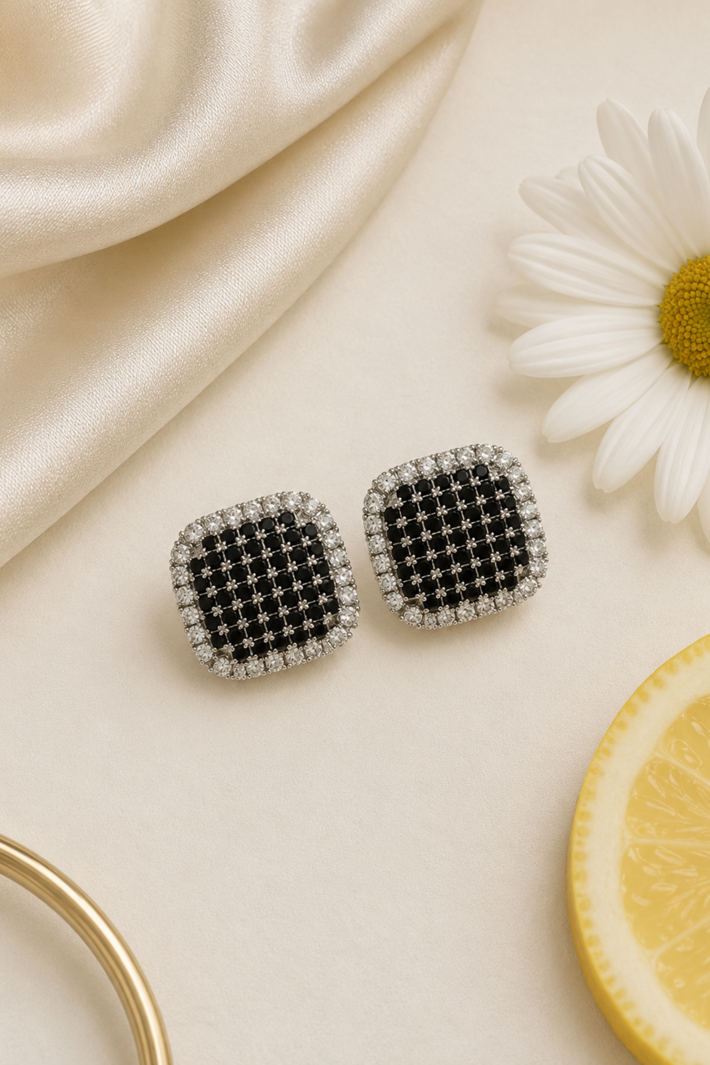 A pair of square stud earrings with a black and silver checkered pattern, surrounded by clear stones, displayed on a cream surface with a white daisy, a slice of lemon, and draped satin fabric.