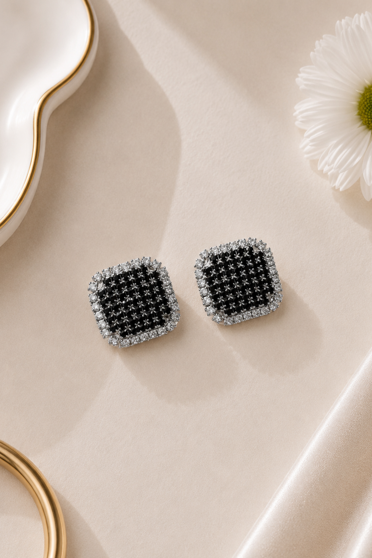 A pair of square stud earrings with a black and silver geometric pattern, styled on a beige satin surface with a white daisy and gold dish props.