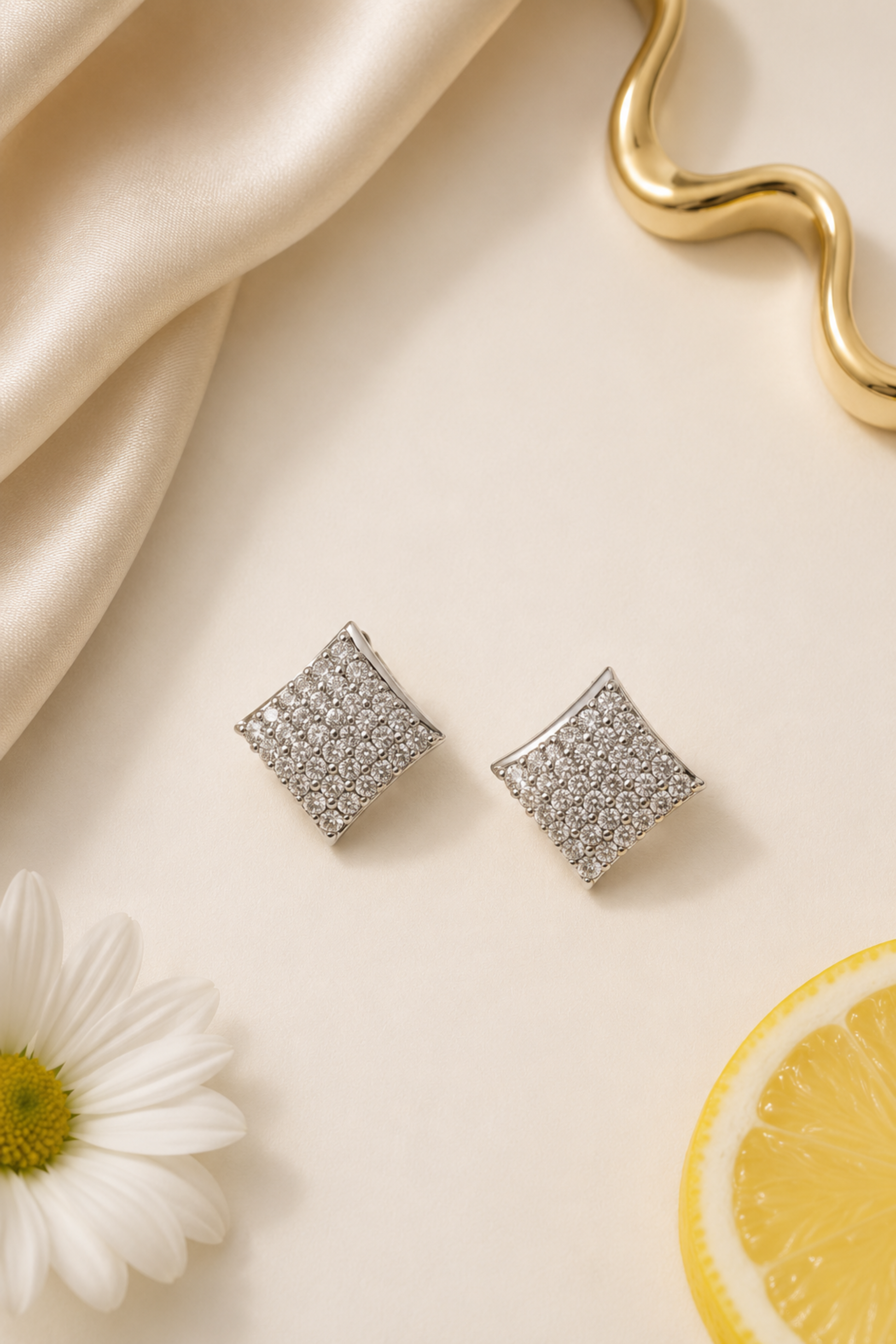 A pair of square stud earrings with a textured, pave-set silver pattern, displayed on a cream-colored surface with a white daisy, a lemon slice, and draped satin fabric.