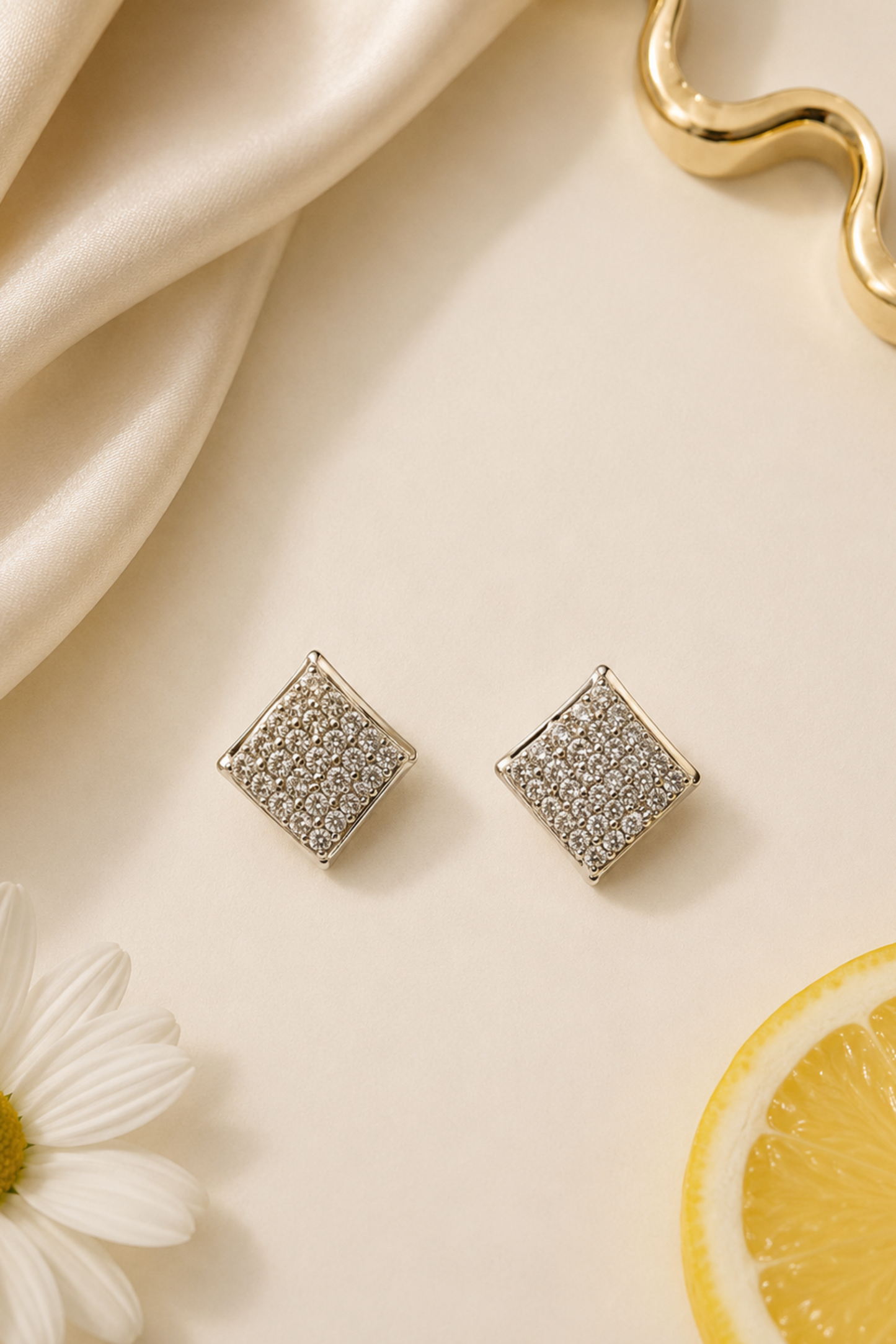 A pair of square stud earrings with a textured, pave-set silver pattern, displayed on a warm cream surface with a draped satin fabric, a white daisy flower, a lemon slice, and a gold wavy object in the background.
