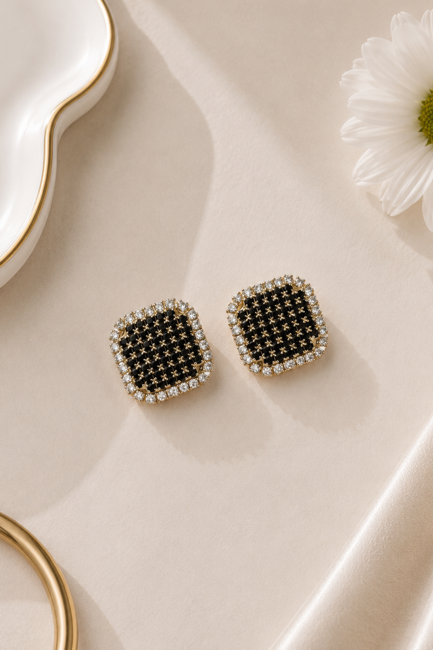 A pair of square stud earrings with black crystal centers and clear crystal borders, displayed on a beige satin fabric next to a white daisy flower and gold jewelry props.