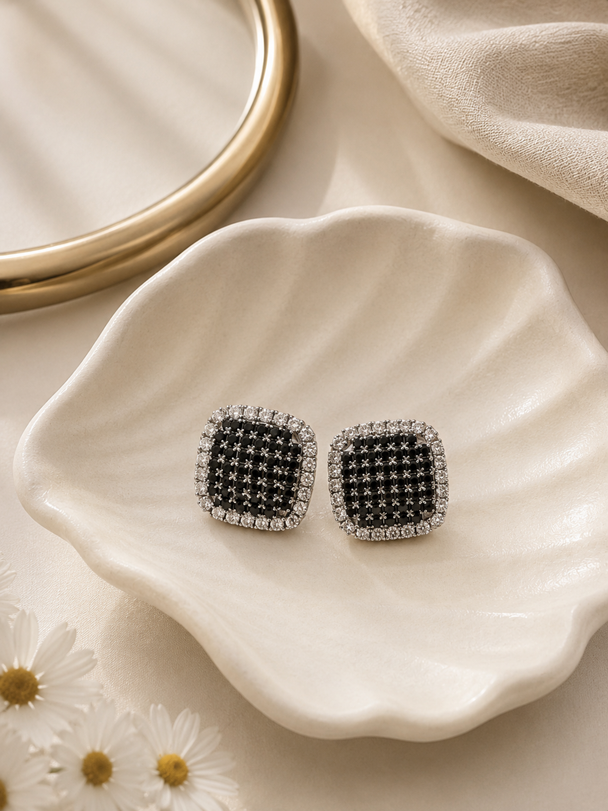 A pair of square stud earrings with black pavé centers and silver crystal borders, displayed on a white ceramic dish with daisies and a gold mirror in the background.