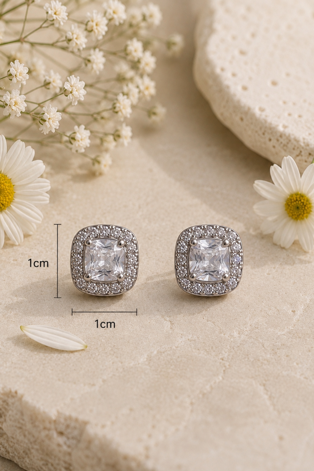 A pair of square stud earrings with clear gemstones in a halo setting, displayed on a textured beige surface with daisies and decorative props. Black measurement lines and text indicate a 1cm width.