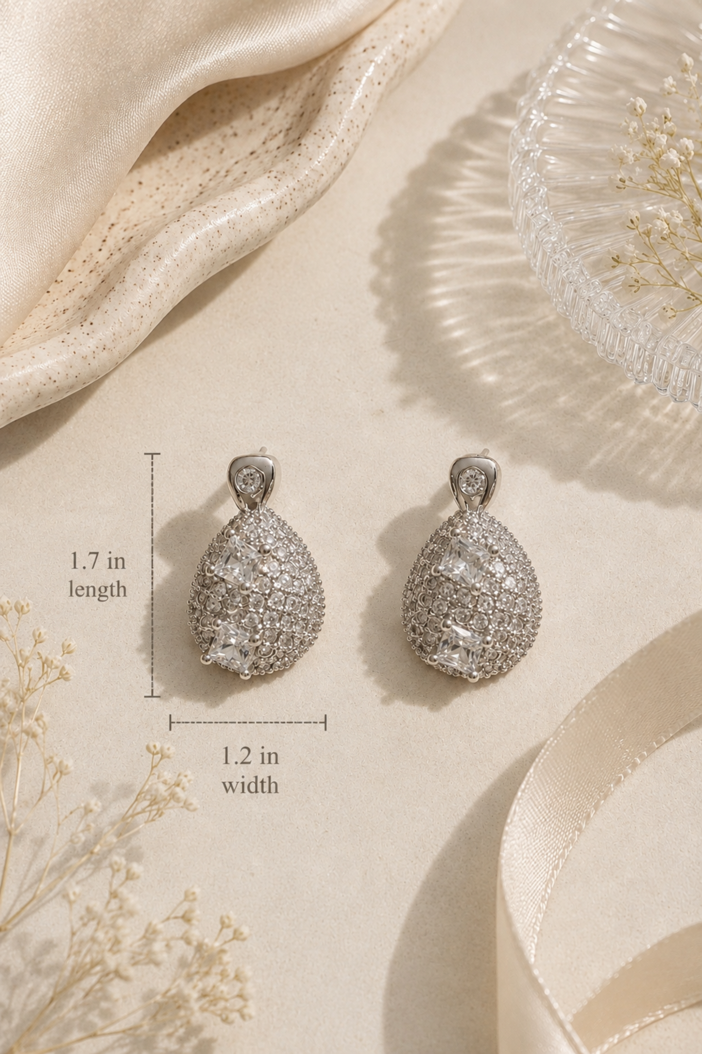 A pair of teardrop-shaped silver earrings covered in clear pavé stones, displayed on a beige fabric background with decorative dried flowers and draped ribbon. Black text overlays indicate the dimensions: 1.