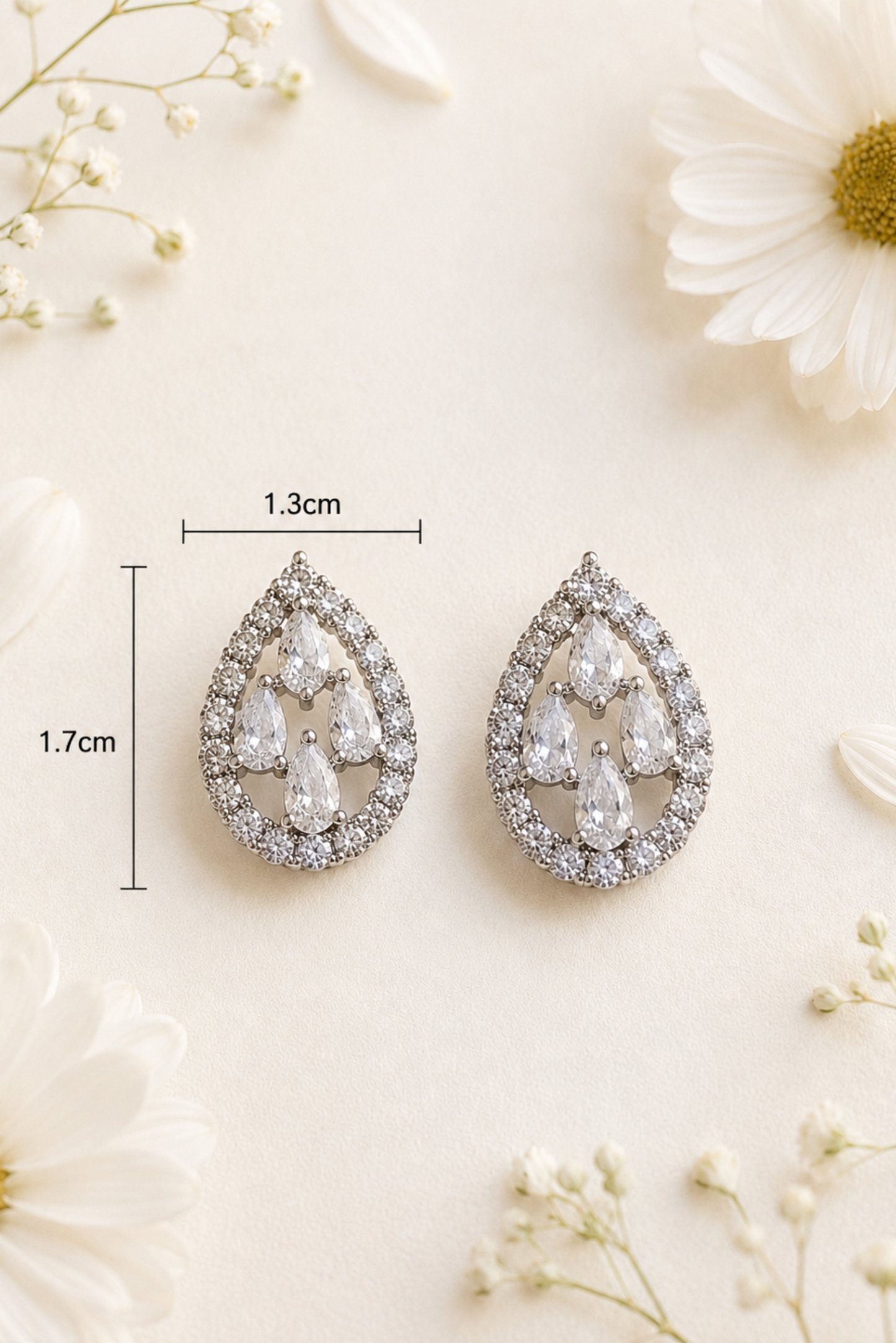 A pair of teardrop-shaped silver earrings with clear gemstones, shown on a light cream background with decorative white flowers and overlaid dimension lines indicating 1.7cm height and 1.