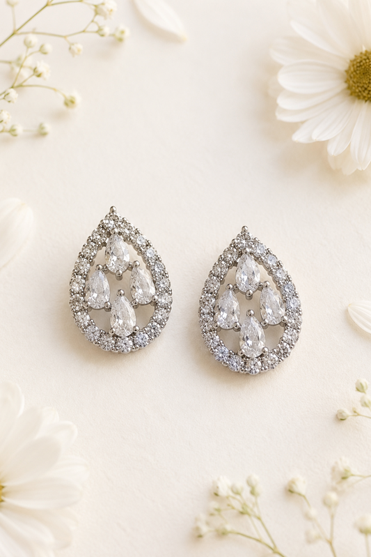 A pair of teardrop-shaped silver stud earrings adorned with clear teardrop and round-cut stones, arranged on a light cream background with white daisies and baby's breath flowers.