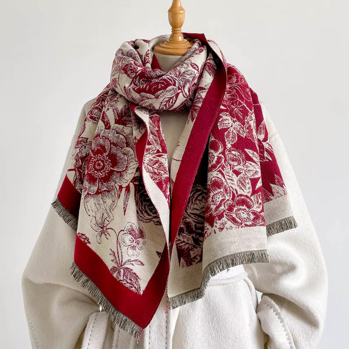 A patterned scarf draped on a cream-colored coat displayed on a mannequin torso. The scarf features a deep red and cream floral print with fringed edges.