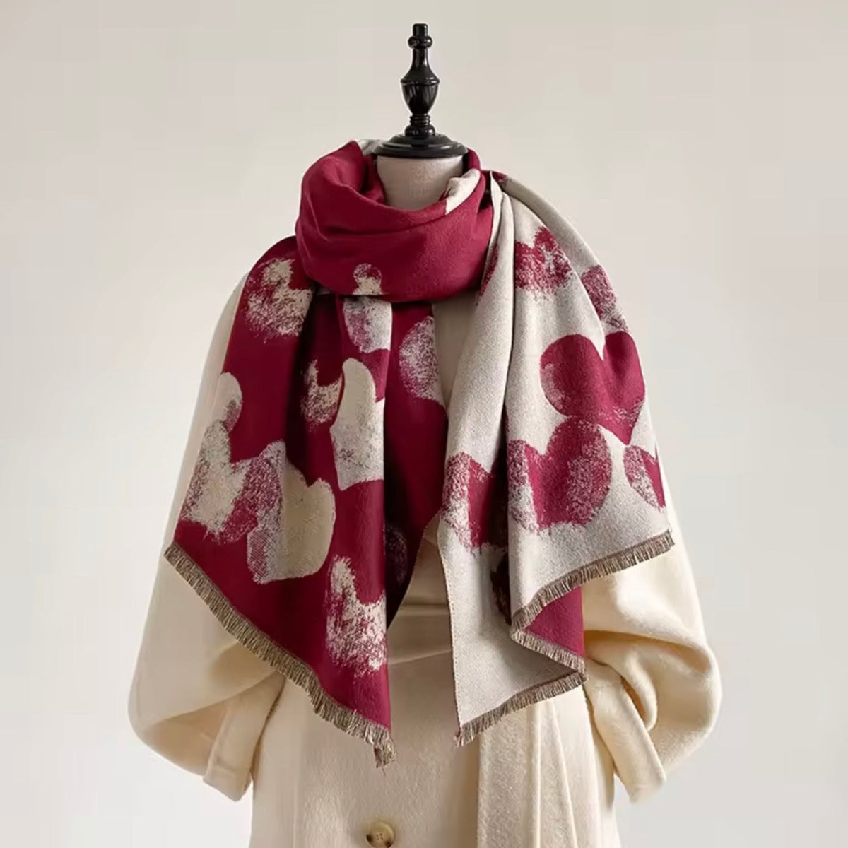 A patterned scarf draped on a cream-colored coat dress form against a light beige background. The scarf features a bold design of deep burgundy abstract shapes on a cream base with fringed edges.