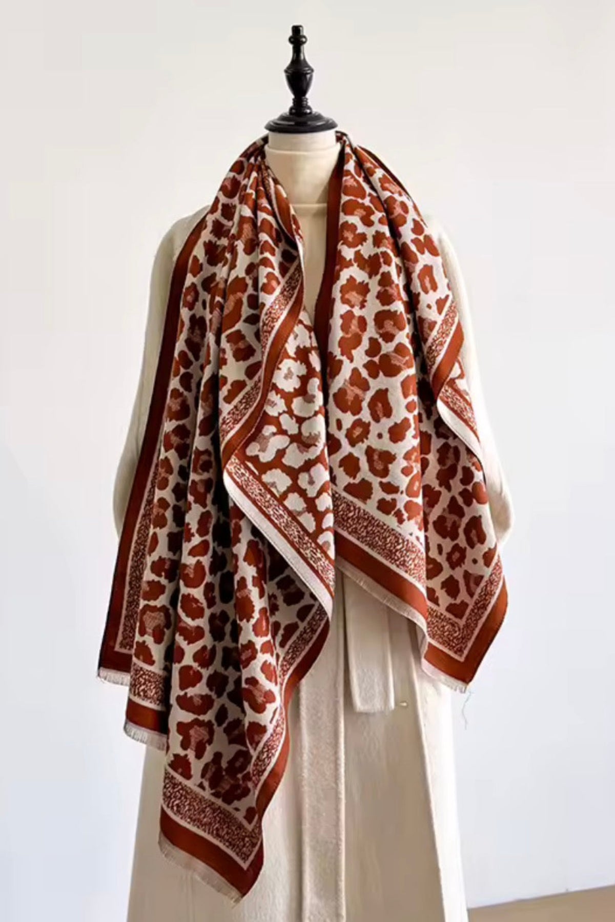 A patterned scarf draped on a cream-colored mannequin dress form against a plain white wall. The scarf features a rust-orange and cream abstract floral pattern with a matching rust-orange border.