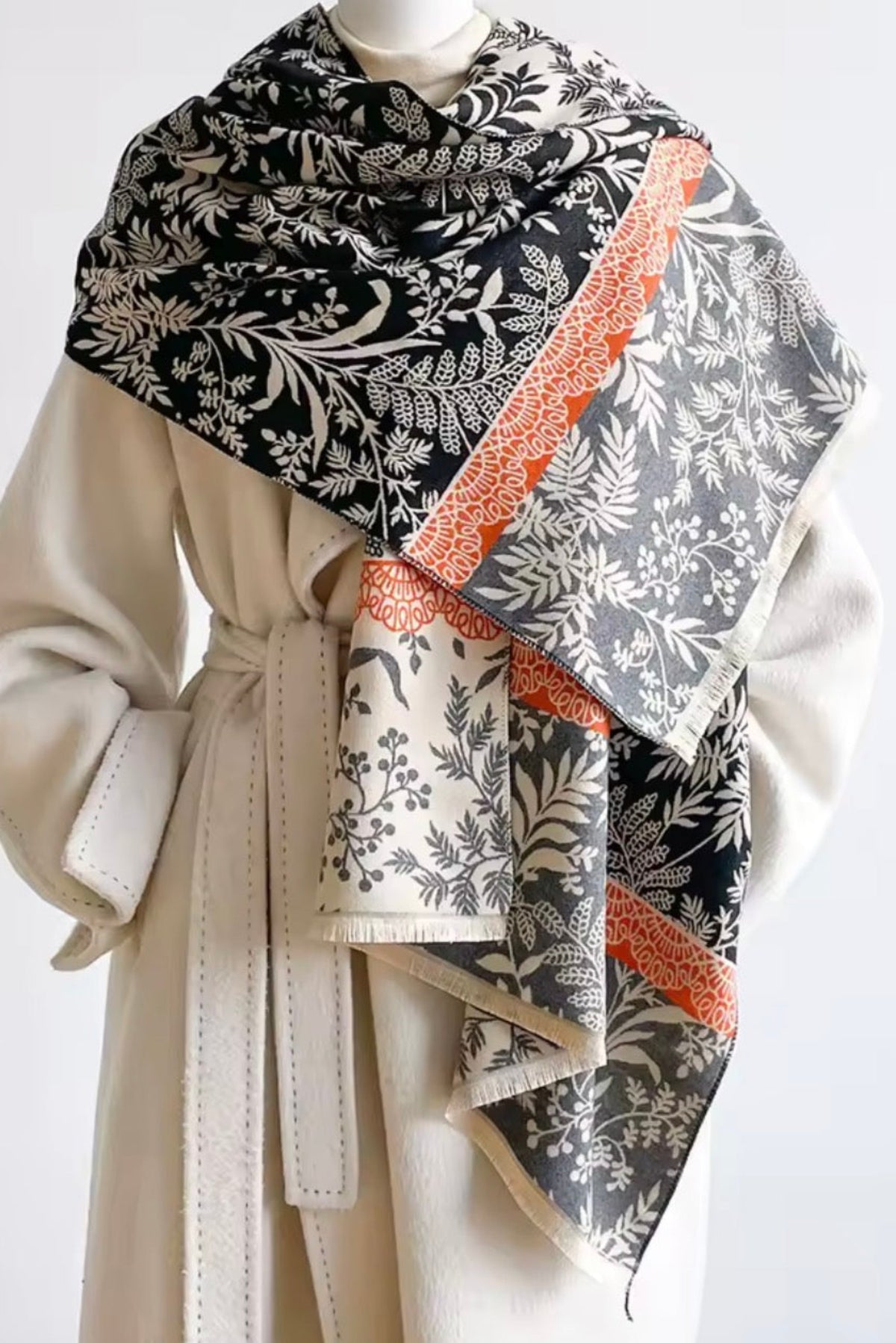 A patterned scarf draped over a cream-colored coat on a mannequin. The scarf features a black and white botanical leaf pattern with orange scalloped borders.
