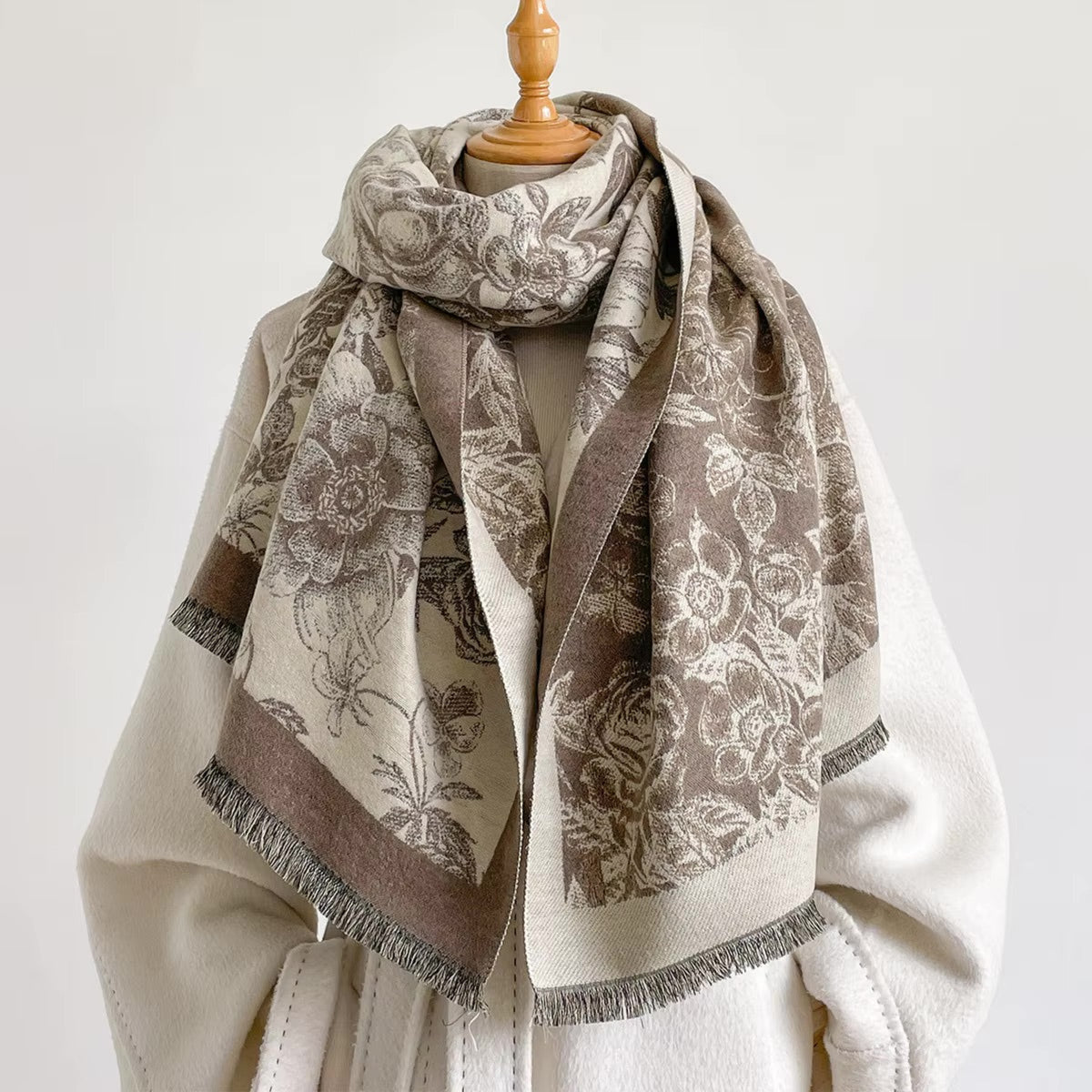 A patterned scarf draped over a cream-colored coat on a mannequin torso. The scarf features a beige and taupe floral damask pattern with fringed edges.