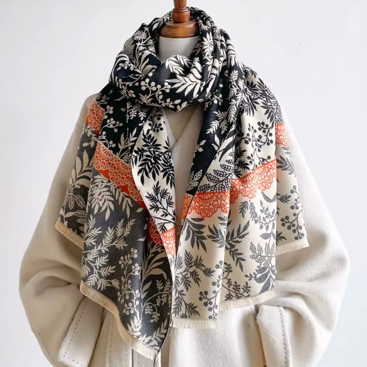 A patterned scarf draped over a cream-colored coat on a mannequin torso. The scarf features botanical leaf prints in black and off-white, accented with orange bands.